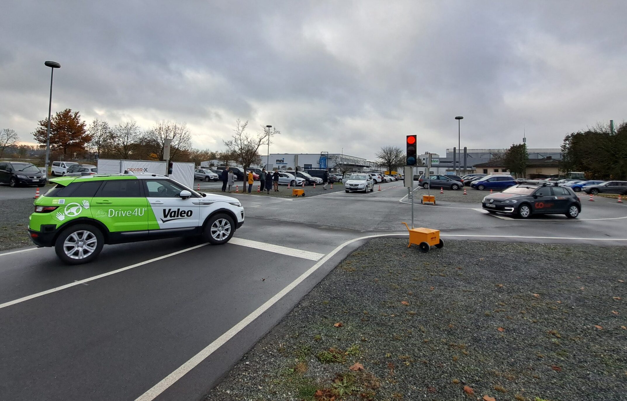 Ein grün-weißes Auto mit der Aufschrift "Valeo Drive4U" wartet an einem bewölkten Tag an einer Ampel auf einem Parkplatz und wirbt für sichere Straßen. Andere Autos parken oder fahren im Hintergrund, während Menschen in der Nähe der geparkten Autos stehen. at Hochschule Coburg