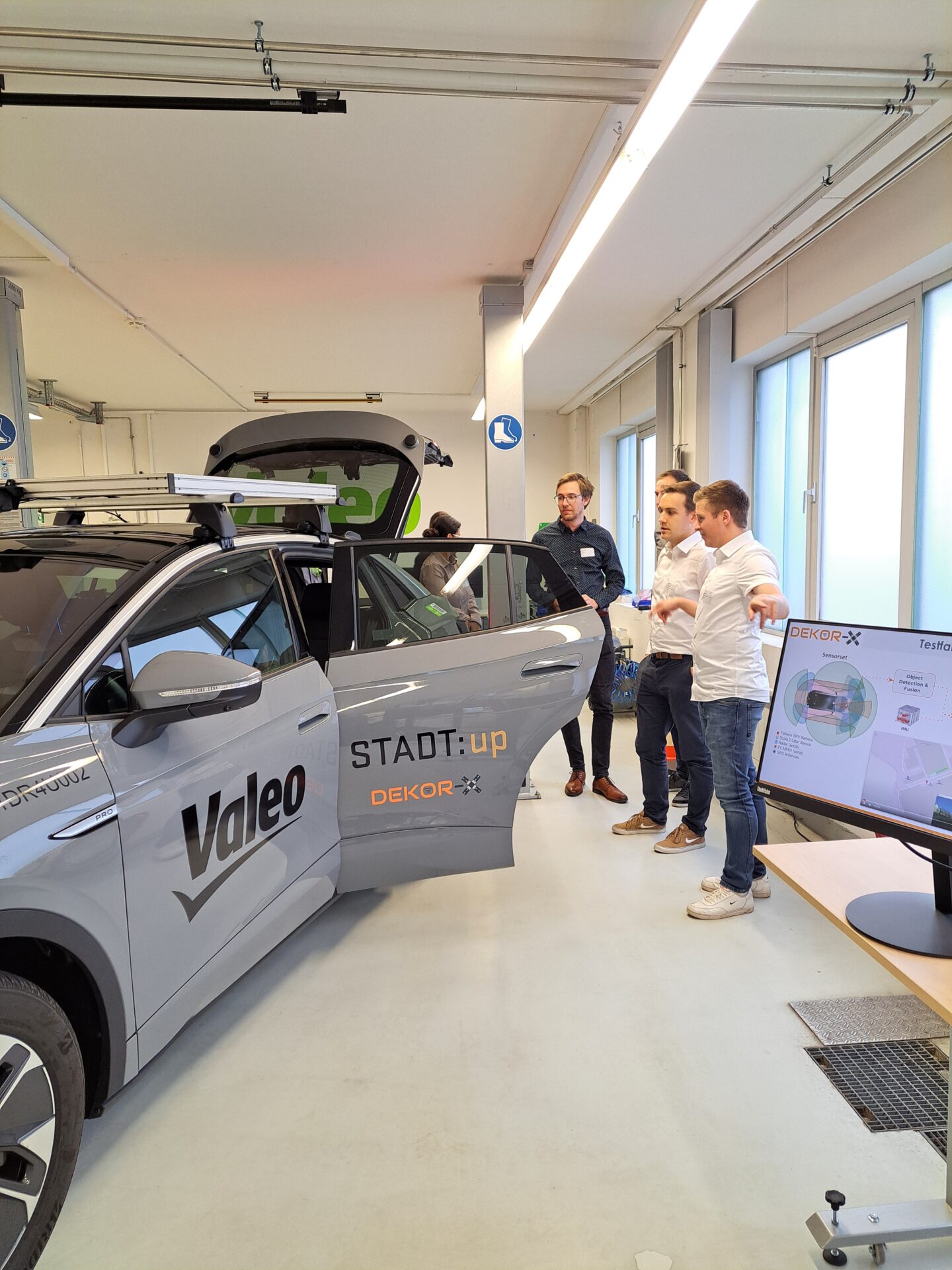 Vier Personen stehen neben einem grauen Auto mit den Aufschriften "Valeo", "STADT:up" und "DEKOR-X" in einem hellen Raum und diskutieren, während eine Person darin sitzt. Auf einem Bildschirm in der Nähe wird eine Präsentation über smarte Kommunikation und Diagramme gezeigt. at Hochschule Coburg