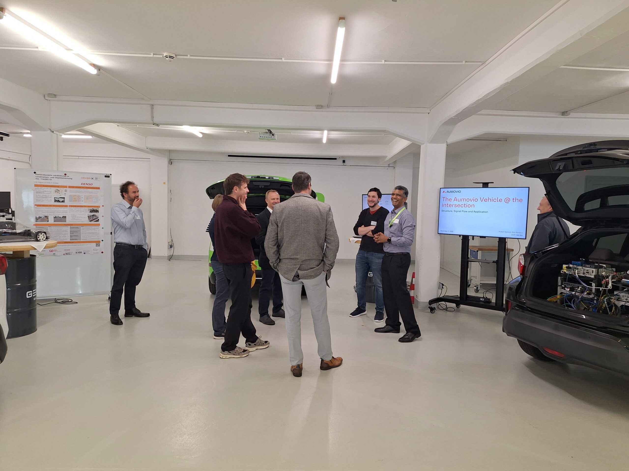 Eine Gruppe von Menschen steht und unterhält sich in einer hellen, modernen Garage mit Autos, DEKOR-X-Plakaten und einer Präsentationsleinwand, auf der "The Autono Vehicle @ the Intersection" zu sehen ist. Einige Teilnehmer beobachten, während eine Person über sichere Straßen spricht. at Hochschule Coburg