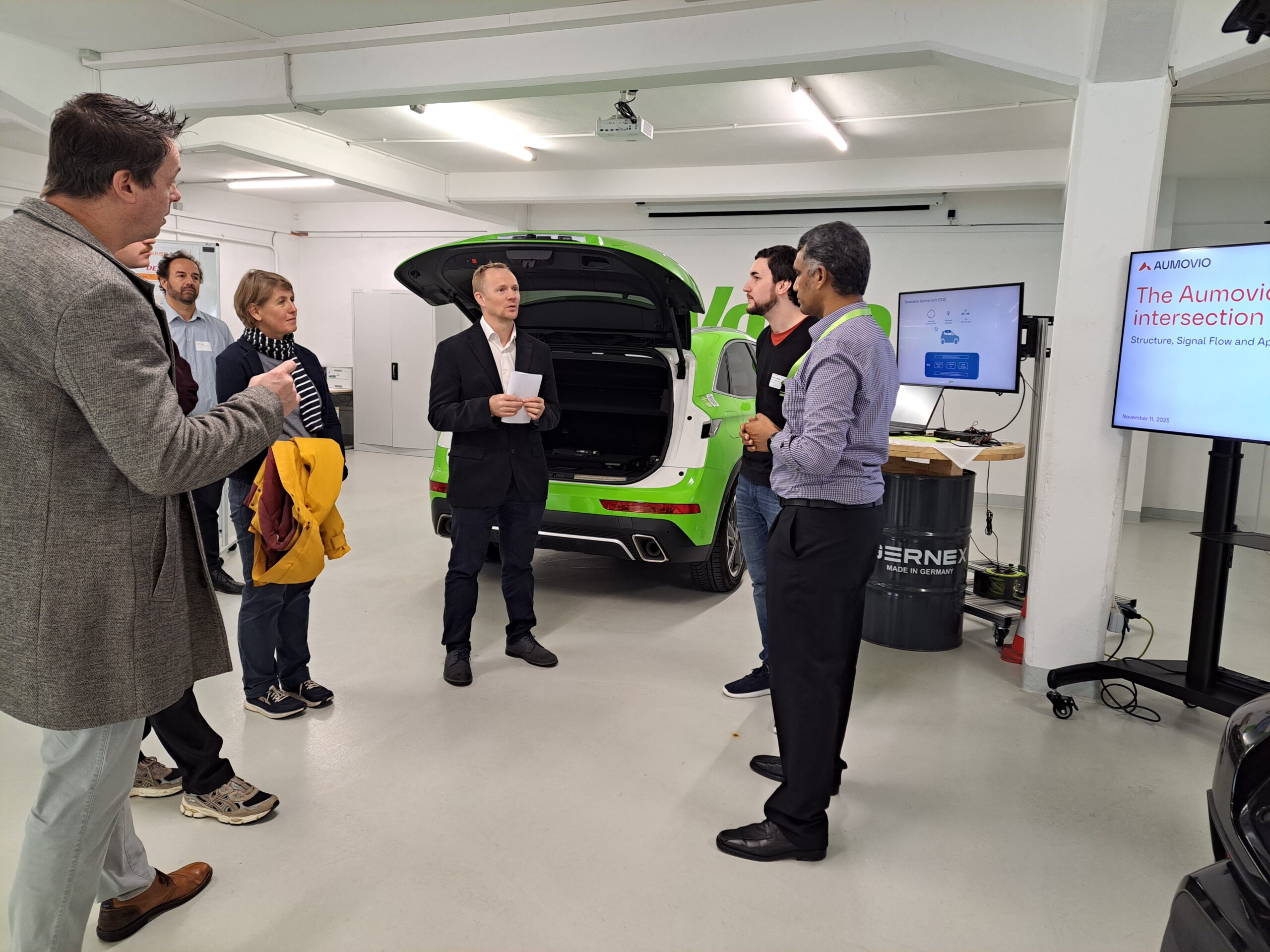 Eine Gruppe von Personen steht in einer modernen Garage um einen offenen Kofferraum herum, während zwei Männer neben einem Bildschirm sprechen, der eine Präsentation mit dem Titel "The Automotive Intersection" mit DEKOR-X-Funktionen für smarte Kommunikation und sichere Straßen zeigt. at Hochschule Coburg