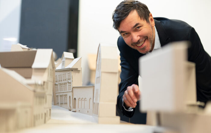 Ein Mann im Anzug lächelt und zeigt auf ein detailliertes Architekturmodell von Gebäuden auf einem Tisch. Er diskutiert über innovative Städte-Designs für die Zukunft als Antwort auf die Klimakrise. at Hochschule Coburg
