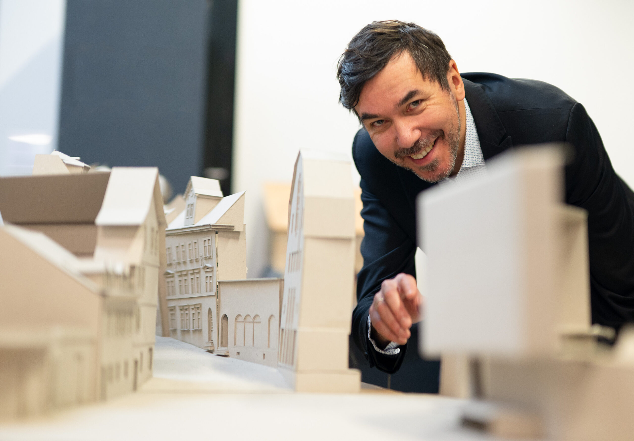 Ein Mann im Anzug lächelt und zeigt auf ein detailliertes Architekturmodell von Gebäuden auf einem Tisch. Er diskutiert über innovative Städte-Designs für die Zukunft als Antwort auf die Klimakrise. at Hochschule Coburg