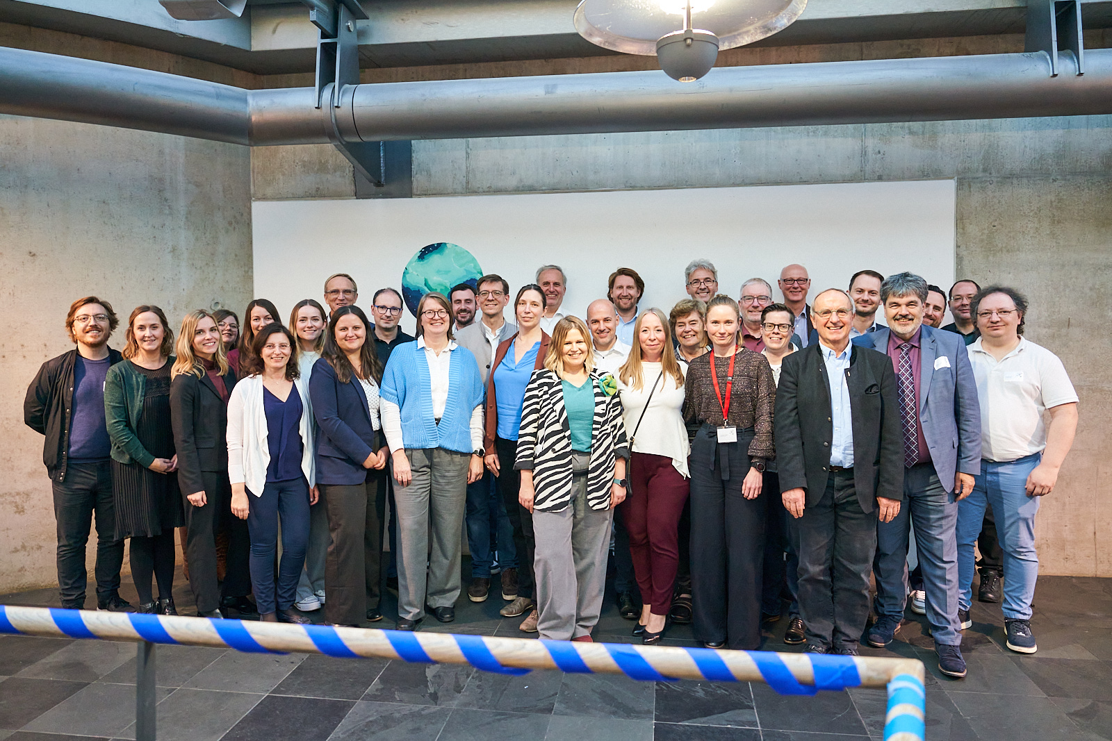 Rund 30 Männer und Frauen posieren in einem Innenraum vor einer weißen Wand mit blau-grünem Rundgemälde für ein Gruppenfoto. Zu sehen ist eine blau-weiße Absperrung, die das Projekt ii.oo auf dem Weg zu digitalen Prüfungen in Bayern begleitet. at Hochschule Coburg