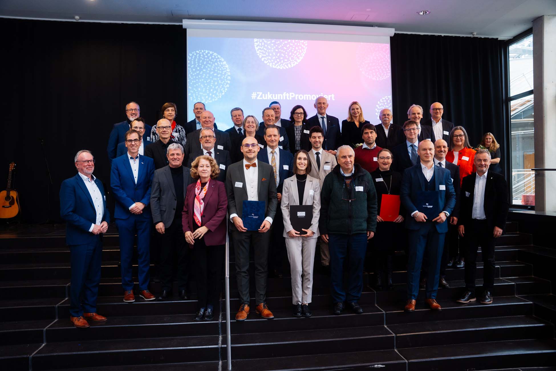 Eine große Gruppe von Absolventen in formeller Kleidung steht in Reihen auf der Bühne, einige halten Urkunden in der Hand. Hinter ihnen zeigt ein Beamer "#ZukunftPromoviert", der die Leistungen der bayerischen Hochschulen und ihrer Promotionszentren hervorhebt. at Hochschule Coburg