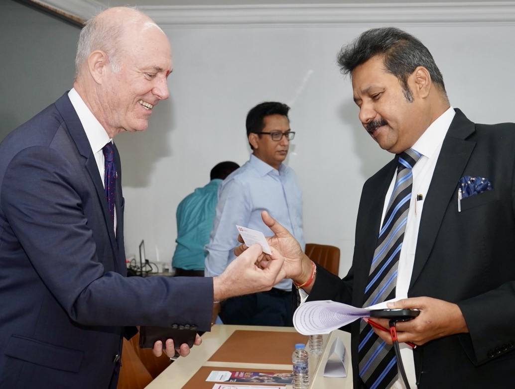 Zwei Männer in Geschäftskleidung, die beide einen Doppel-MBA anstreben, lächeln und tauschen während eines beruflichen Treffens über einen Tisch hinweg eine Visitenkarte aus, während eine dritte Person im Hintergrund steht. at Hochschule Coburg