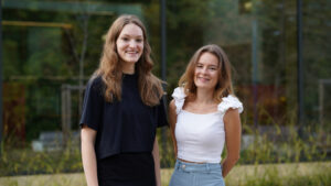 Two young women are standing side by side outdoors, smiling at the camera. One wears a black top and has loose hair, while the other wears a white top with ruffled sleeves and blue pants. Greenery is visible in the background. at Hochschule Coburg