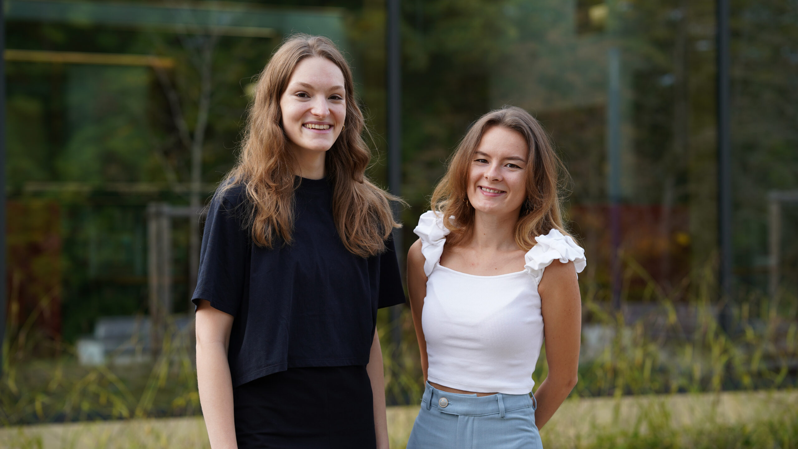 Zwei junge Frauen stehen im Freien und lächeln in die Kamera. Die eine trägt ein schwarzes Oberteil, die andere ein weißes Oberteil mit gerüschten Ärmeln und eine hellblaue Hose. Im Hintergrund sind grünes Laub und ein Glasgebäude zu sehen. at Hochschule Coburg