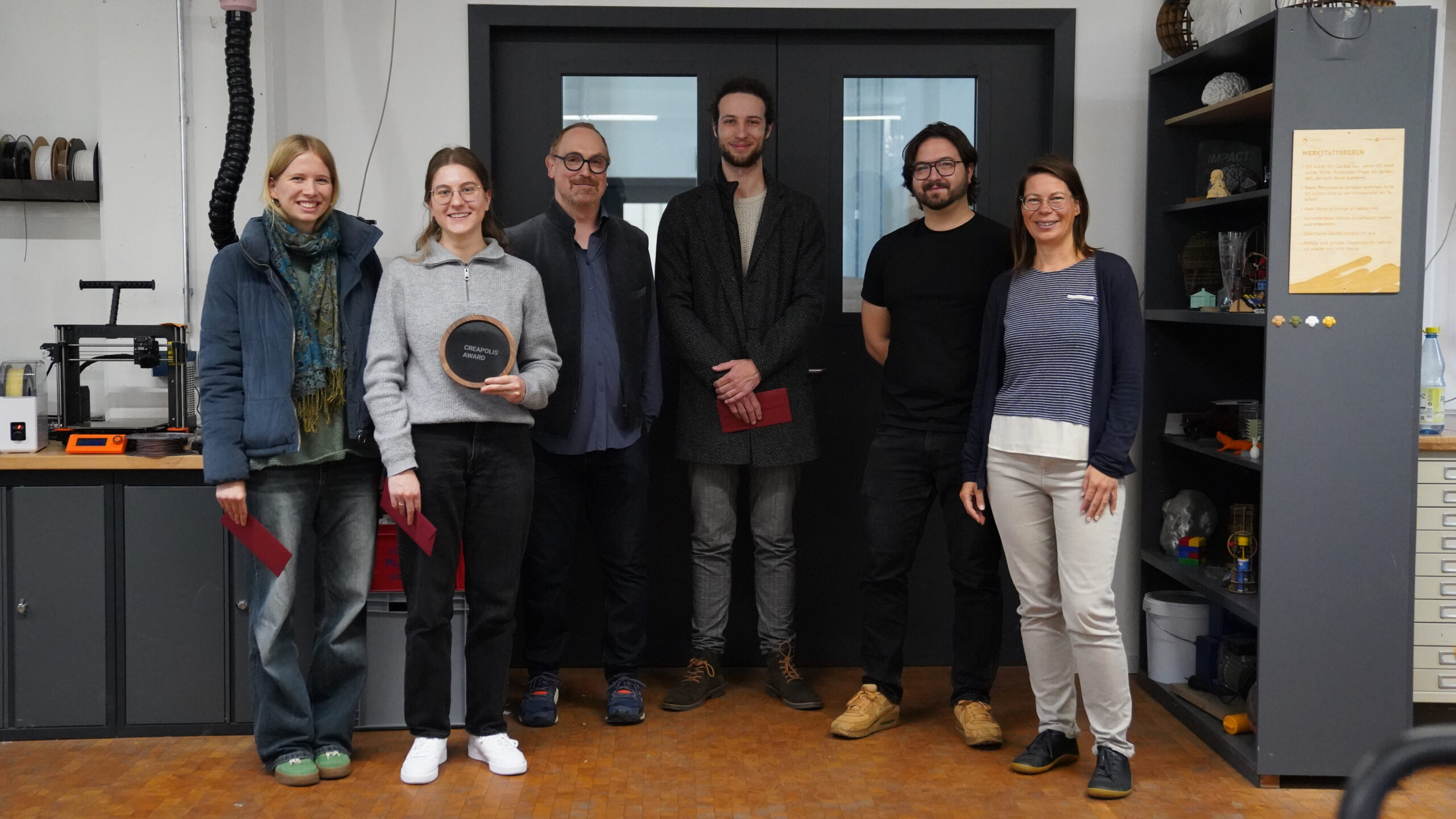 Sechs Personen stehen in einem Innenraum in einer Werkstatt oder einem Labor und lächeln in die Kamera. Einige halten rote Umschläge in der Hand, während eine Person stolz eine schwarze runde CREAPOLIS-Plakette zeigt. Um sie herum sind Regale, Schränke und 3D-Designmodelle zu sehen. at Hochschule Coburg