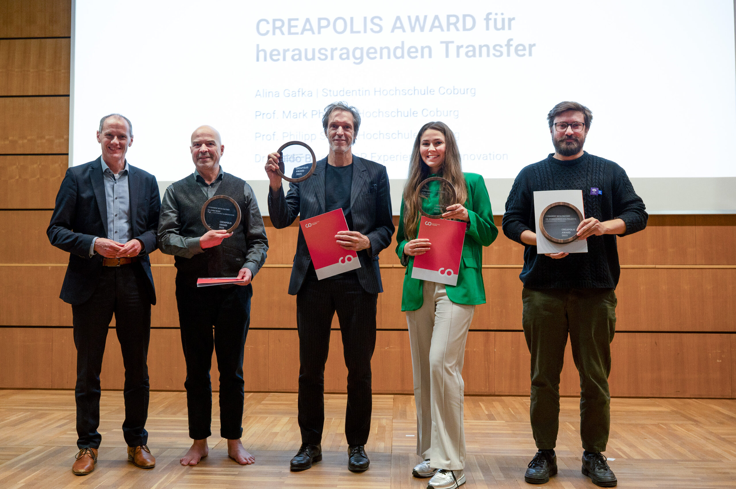 Fünf Personen stehen nebeneinander auf einer hölzernen Bühne beim Dies academicus 2025 der Hochschule, halten Preise und Urkunden in der Hand und lächeln für ein Foto. Hinter ihnen steht auf einer Projektion "CREAPOLIS AWARD für herausragenden Transfer" und zählt mehrere Namen auf. at Hochschule Coburg