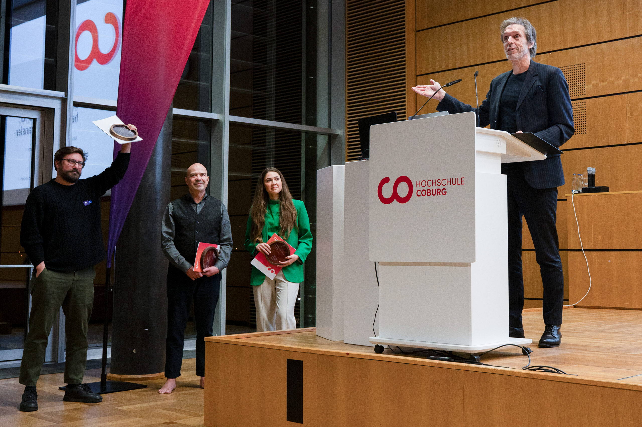 Vier Personen nehmen an einer Veranstaltung in einer modernen Halle teil. Einer spricht an einem Podium mit der Aufschrift "Hochschule Coburg", während die anderen in der Nähe stehen, lächeln und Gegenstände in der Hand halten - einer hebt den Hut zur Begrüßung - beim CREAPOLIS-Award, mit dem Bürokonzepte der Zukunft ausgezeichnet werden. at Hochschule Coburg