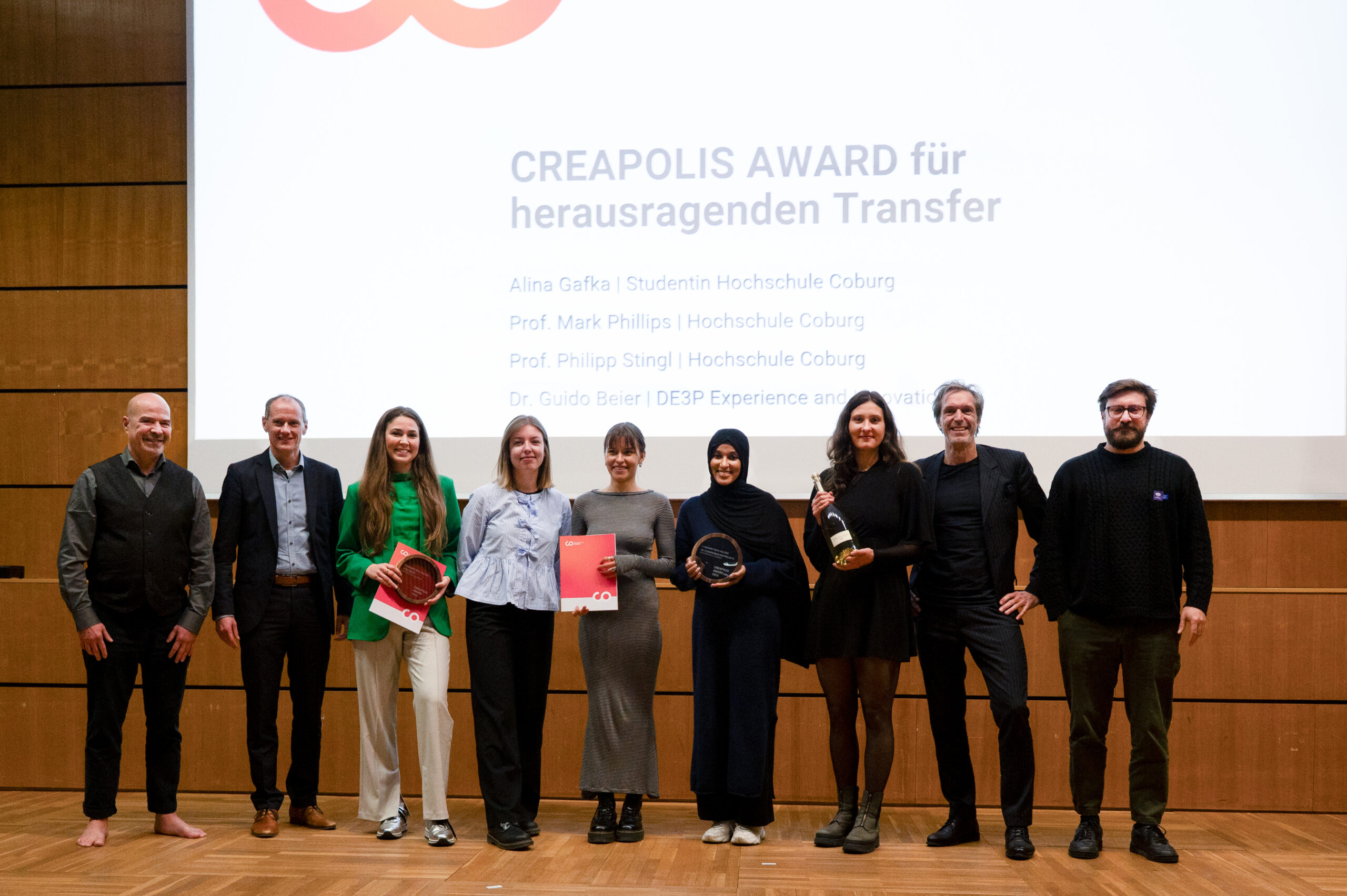 Acht Personen stehen auf einer hölzernen Bühne, halten Preise und Urkunden in der Hand und lächeln für ein Gruppenfoto vor einem Bildschirm mit der Aufschrift "CREAPOLIS-Award für herausragenden Transfer - Bürokonzepte der Zukunft". at Hochschule Coburg