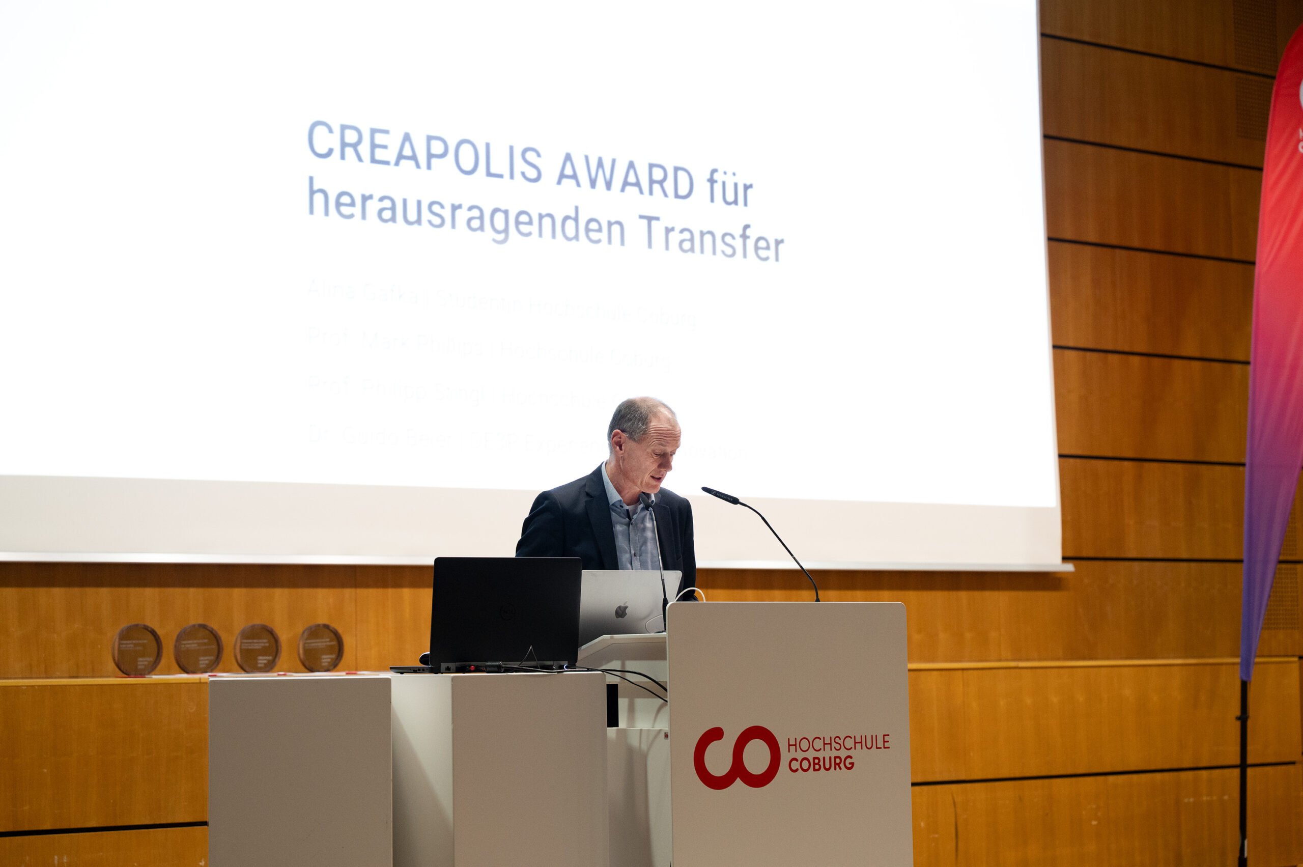 Ein Mann steht an einem Podium mit einem Mikrofon und spricht bei der Veranstaltung "CREAPOLIS-Award für herausragenden Transfer" an der Hochschule Coburg, bei der innovative Bürokonzepte der Zukunft ausgezeichnet werden. Auf einem Tisch in der Nähe sind drei Preisplaketten zu sehen. at Hochschule Coburg
