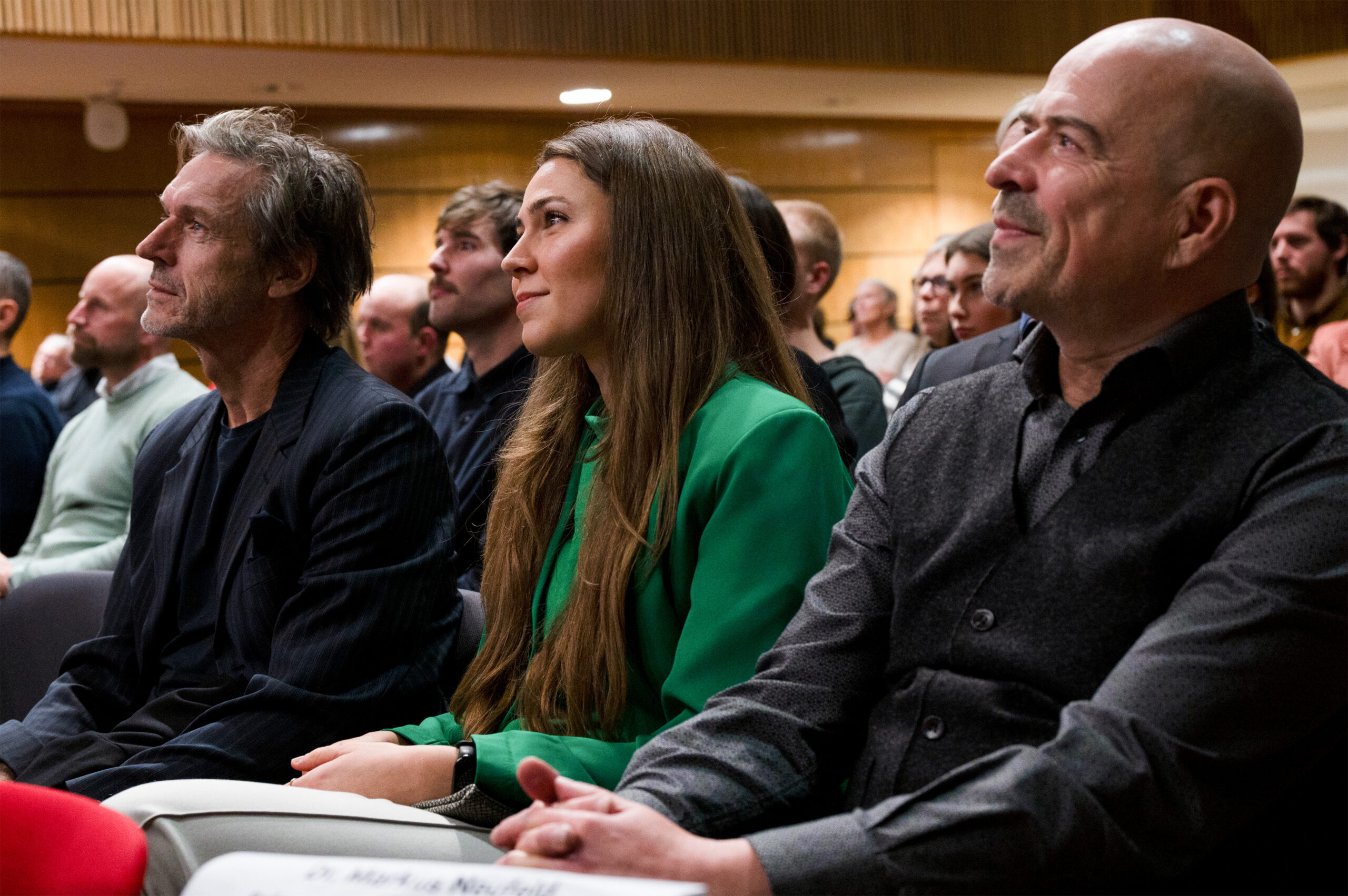 Eine Gruppe von Menschen sitzt aufmerksam im Publikum und konzentriert sich auf etwas, das sie nicht sehen können. Bei der CREAPOLIS-Preisverleihung scheinen ein Mann in einer dunklen Jacke, eine Frau in einer grünen Bluse und ein weiterer Mann in einer grauen Weste ganz bei der Sache zu sein. at Hochschule Coburg