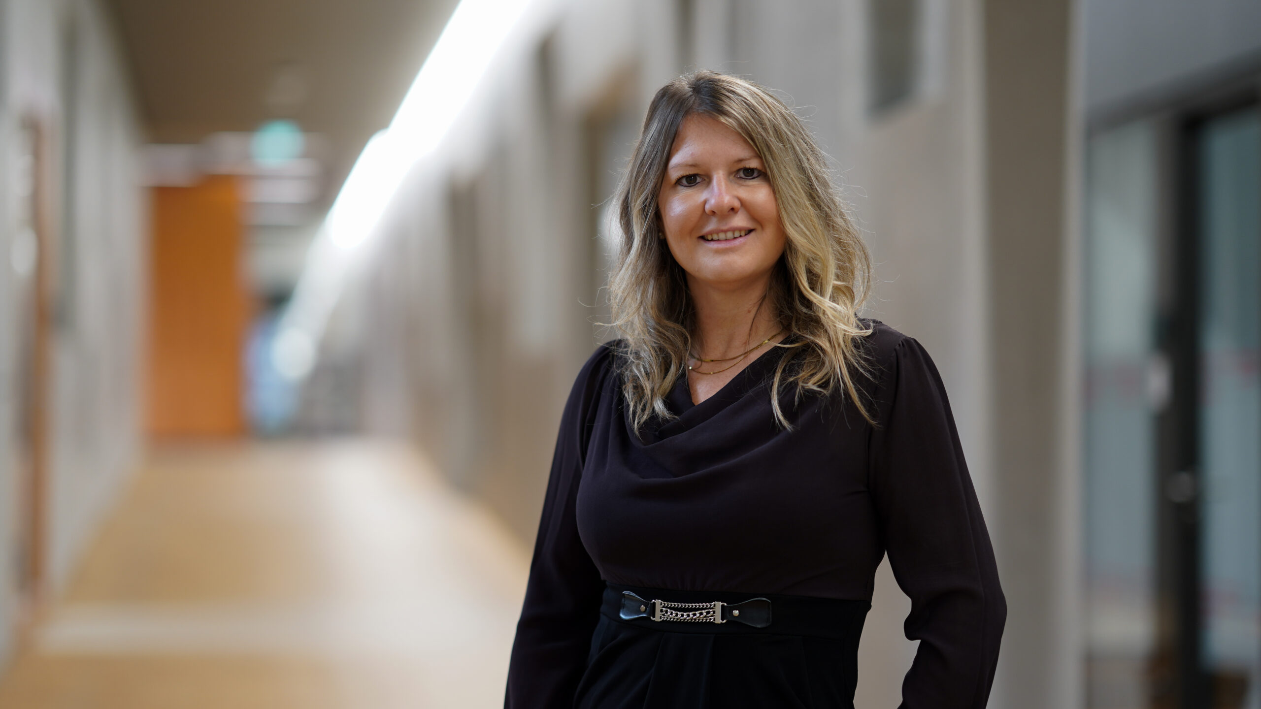 Eine Frau mit langen blonden Haaren und einem schwarzen Kleid steht lächelnd in einem hell erleuchteten Flur mit verschwommenen Wänden und Türen im Hintergrund. at Hochschule Coburg