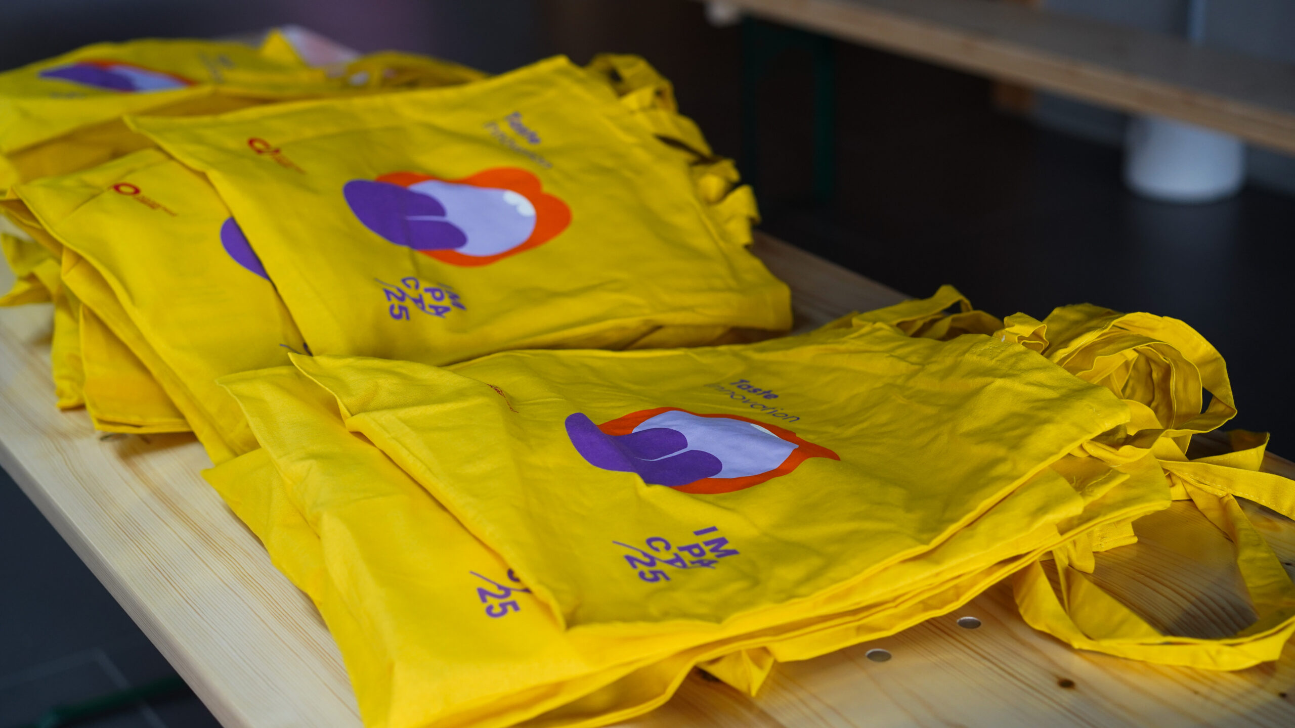 Ein Stapel leuchtend gelber Tragetaschen mit farbenfrohen abstrakten Motiven und einem Text, der Kreativität und Innovation anpreist, liegt auf einem Holztisch bereit zur Verteilung. at Hochschule Coburg