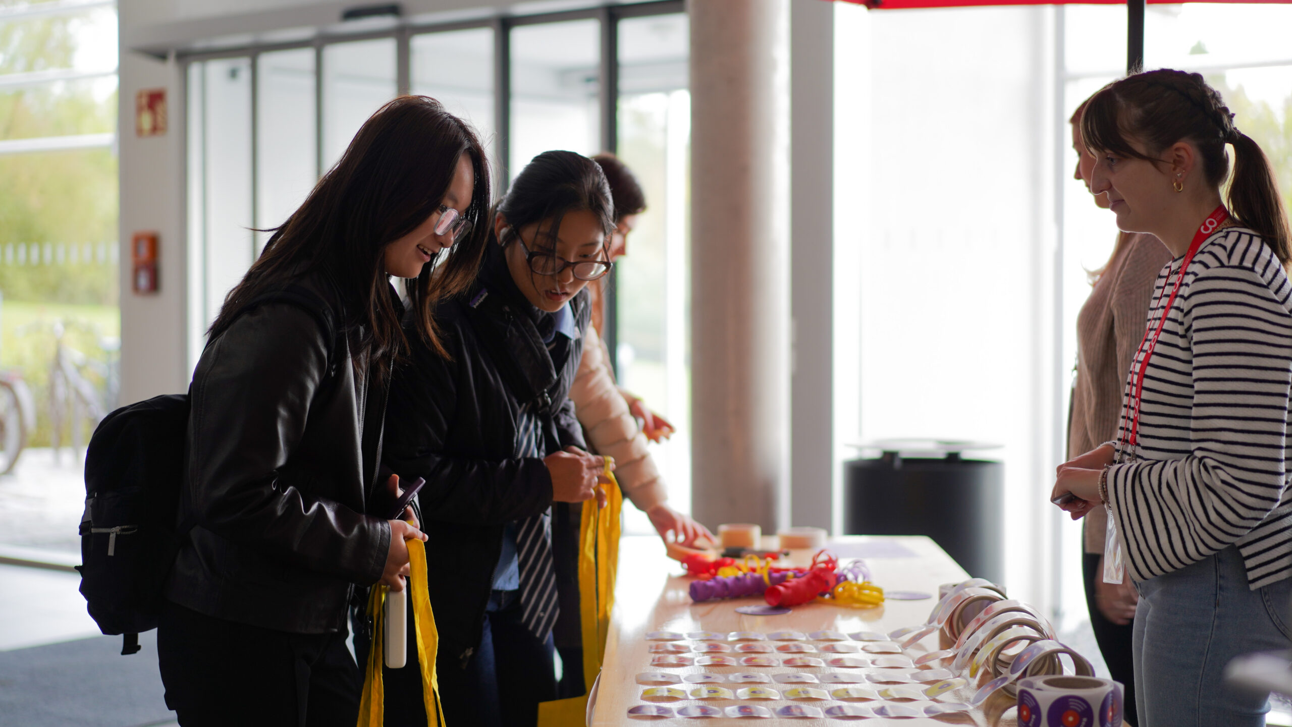 Drei Personen demonstrieren Teamgeist, während sie mit gelben Tragetaschen einen Tisch mit Knöpfen und Snacks untersuchen, während eine Frau auf der anderen Seite ihnen in einem gut beleuchteten Innenraum hilft. at Hochschule Coburg