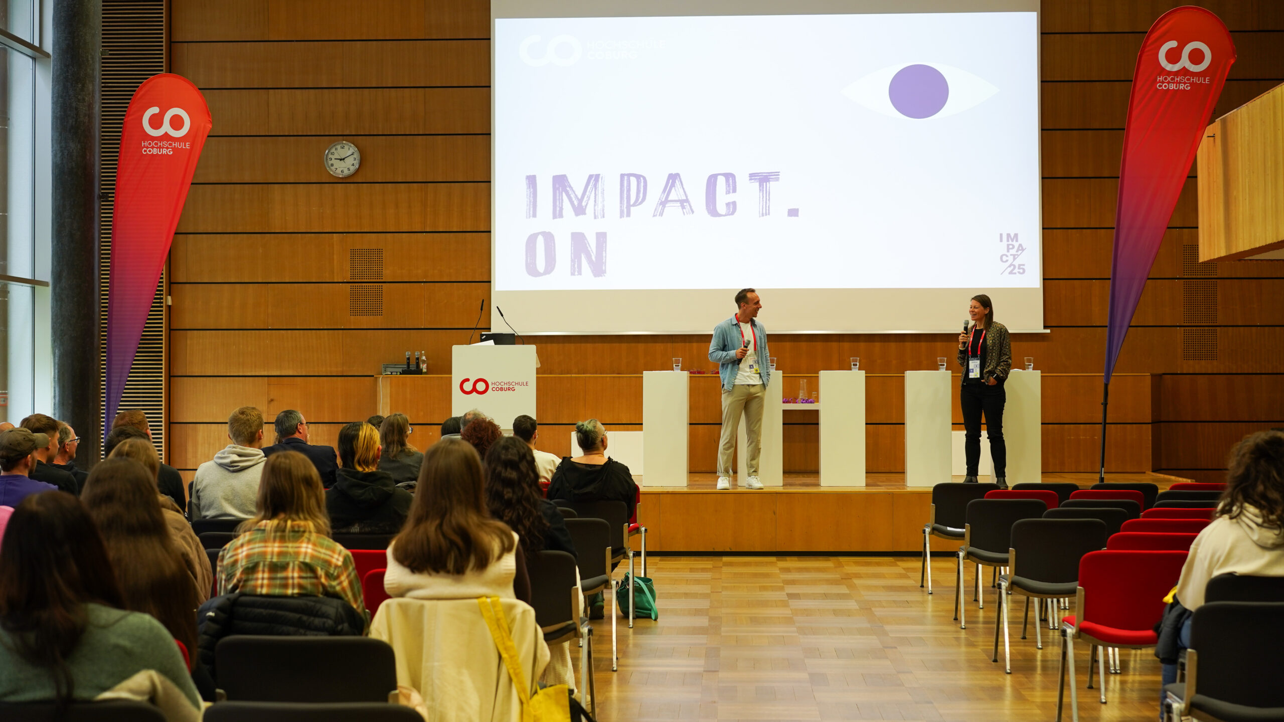 Ein Konferenzraum mit sitzenden Teilnehmern blickt auf eine Bühne, auf der zwei Redner neben einem Bildschirm stehen, auf dem "IMPACT. ON". Das moderne Ambiente mit Holzwänden und rot akzentuierten Bannern fördert den Teamgeist und beflügelt die Innovation unter den Teilnehmern. at Hochschule Coburg