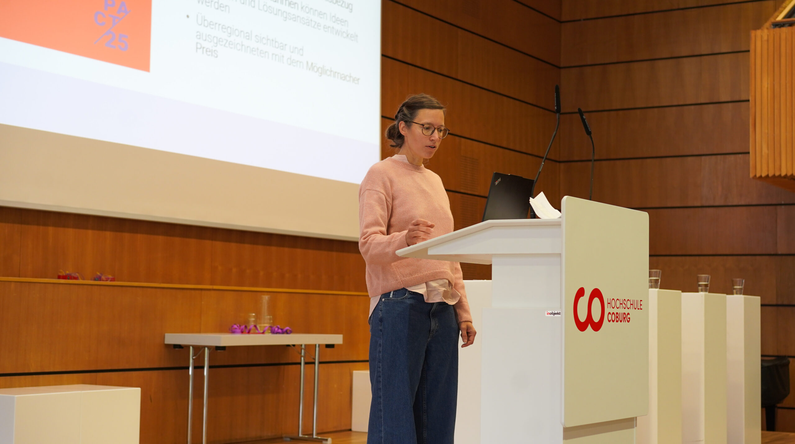 Eine Frau mit Brille, rosa Pullover und blauer Jeans steht an einem weißen Podium und hält in einem holzgetäfelten Hörsaal einen Vortrag. Die Kulisse mit dem Logo der Hochschule Coburg und der Leinwand unterstreicht die Innovation im akademischen Umfeld. at Hochschule Coburg