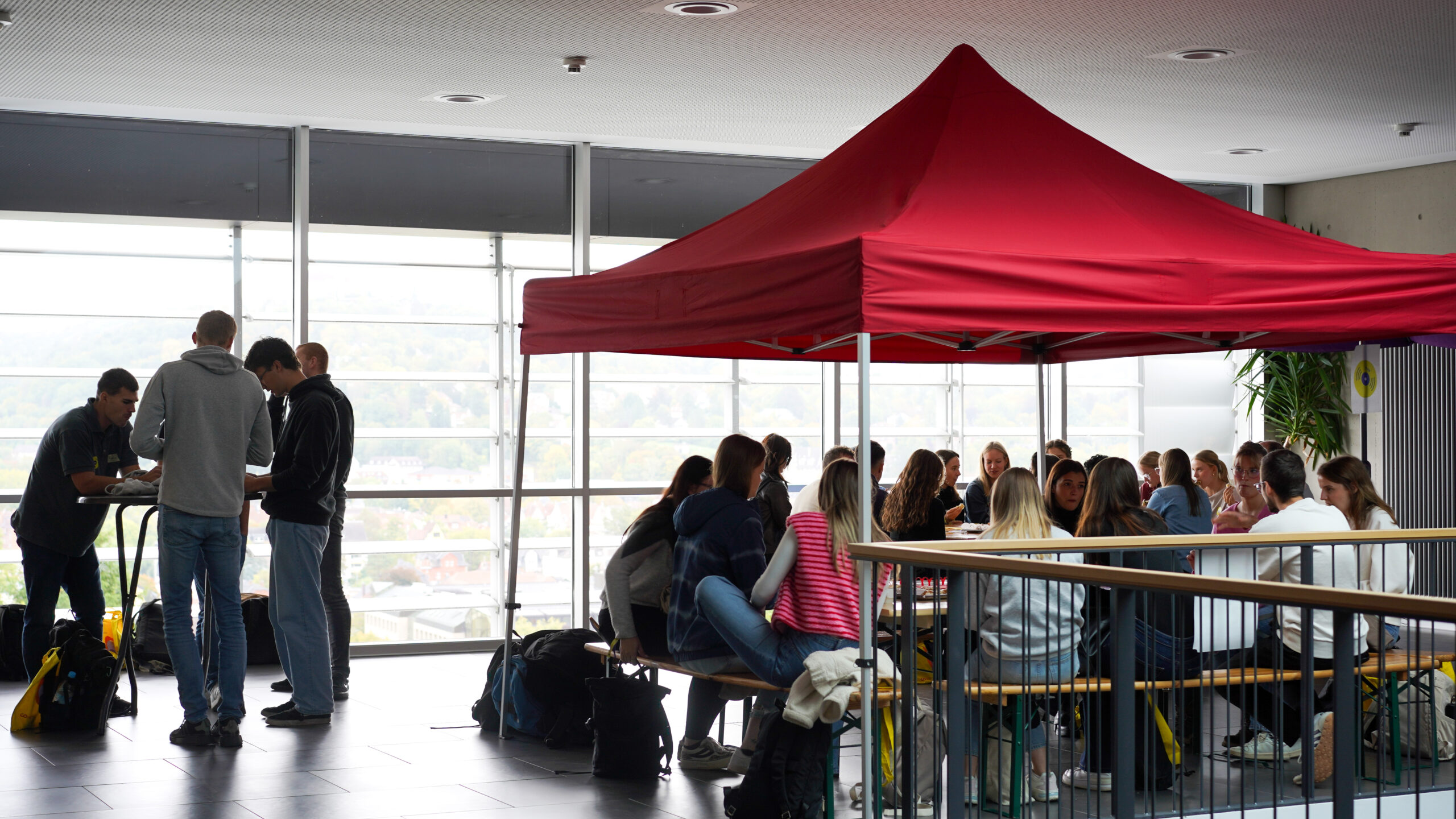 Eine Gruppe von Menschen sitzt und unterhält sich unter einem roten Baldachin, während eine kleinere Gruppe in der Nähe plaudert - große Fenster lassen natürliches Licht herein und schaffen einen inspirierenden Raum, der Teamgeist und Kreativität fördert. at Hochschule Coburg