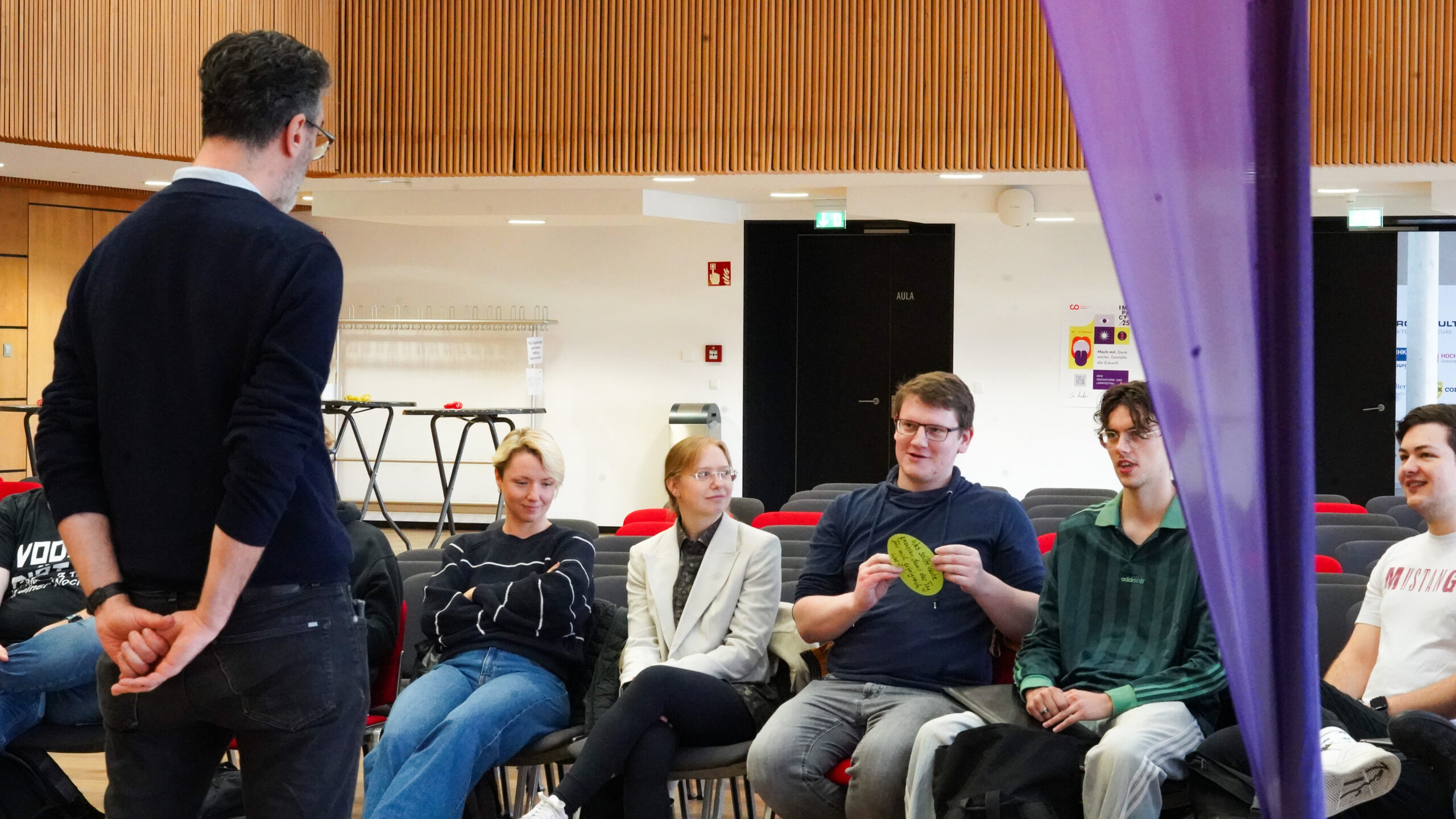 Eine Gruppe von Menschen sitzt in einem Halbkreis von Stühlen in einem Innenraum und hört einem Mann zu, der vor ihnen steht. Eine sitzende Person hält ein gelbes Objekt hoch, das Kreativität und Teamgeist in dem holzgetäfelten Raum mit leeren Stühlen im Hintergrund fördert. at Hochschule Coburg