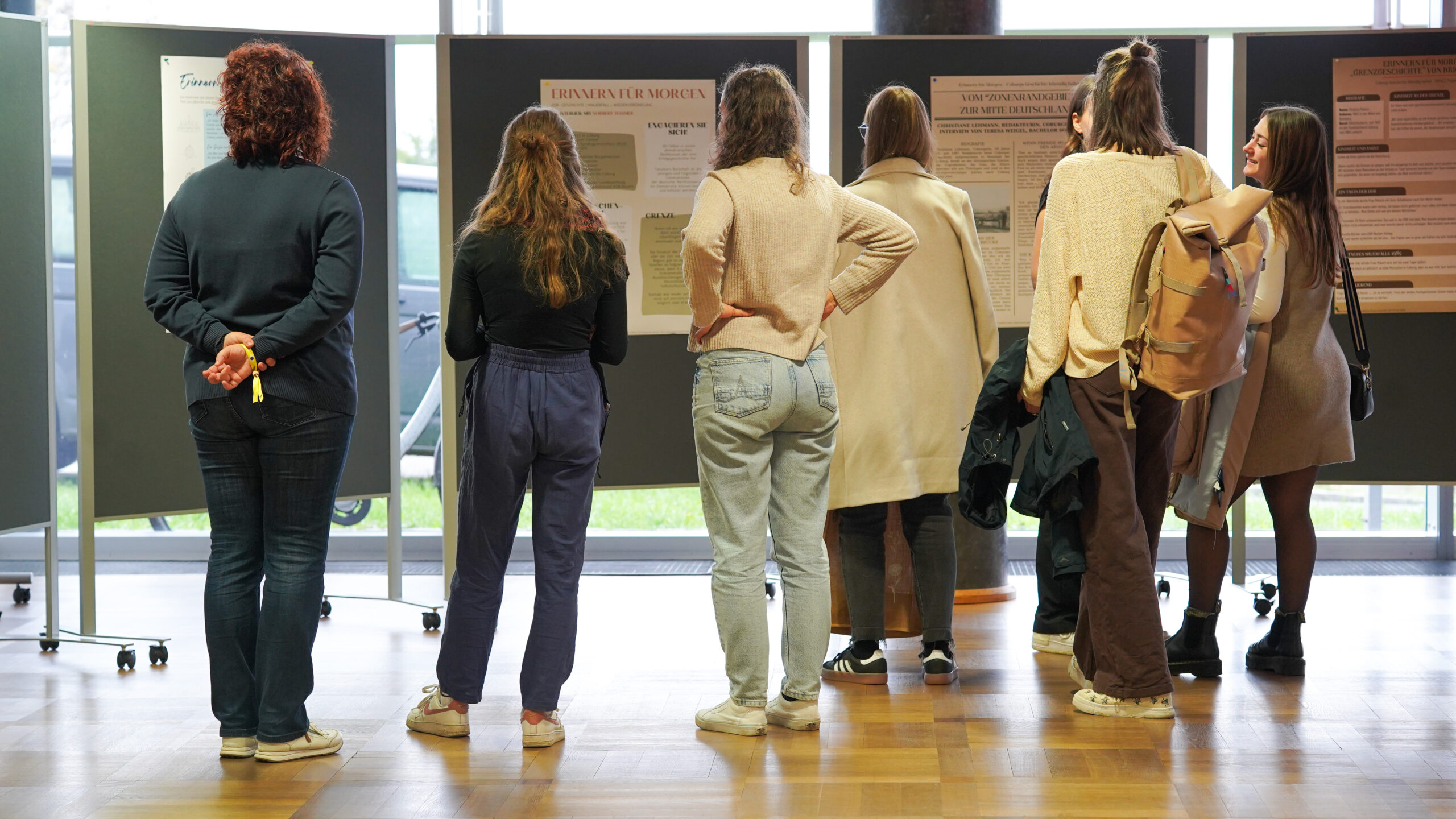 Eine Gruppe von Menschen steht in einem Innenraum vor Informationsplakaten, die auf Stellwänden angebracht sind. Die meisten sind leger gekleidet und scheinen die Plakate zu lesen oder darüber zu diskutieren, was den Teamgeist und die Kreativität in einem Raum mit Holzfußboden und großen Fenstern fördert. at Hochschule Coburg