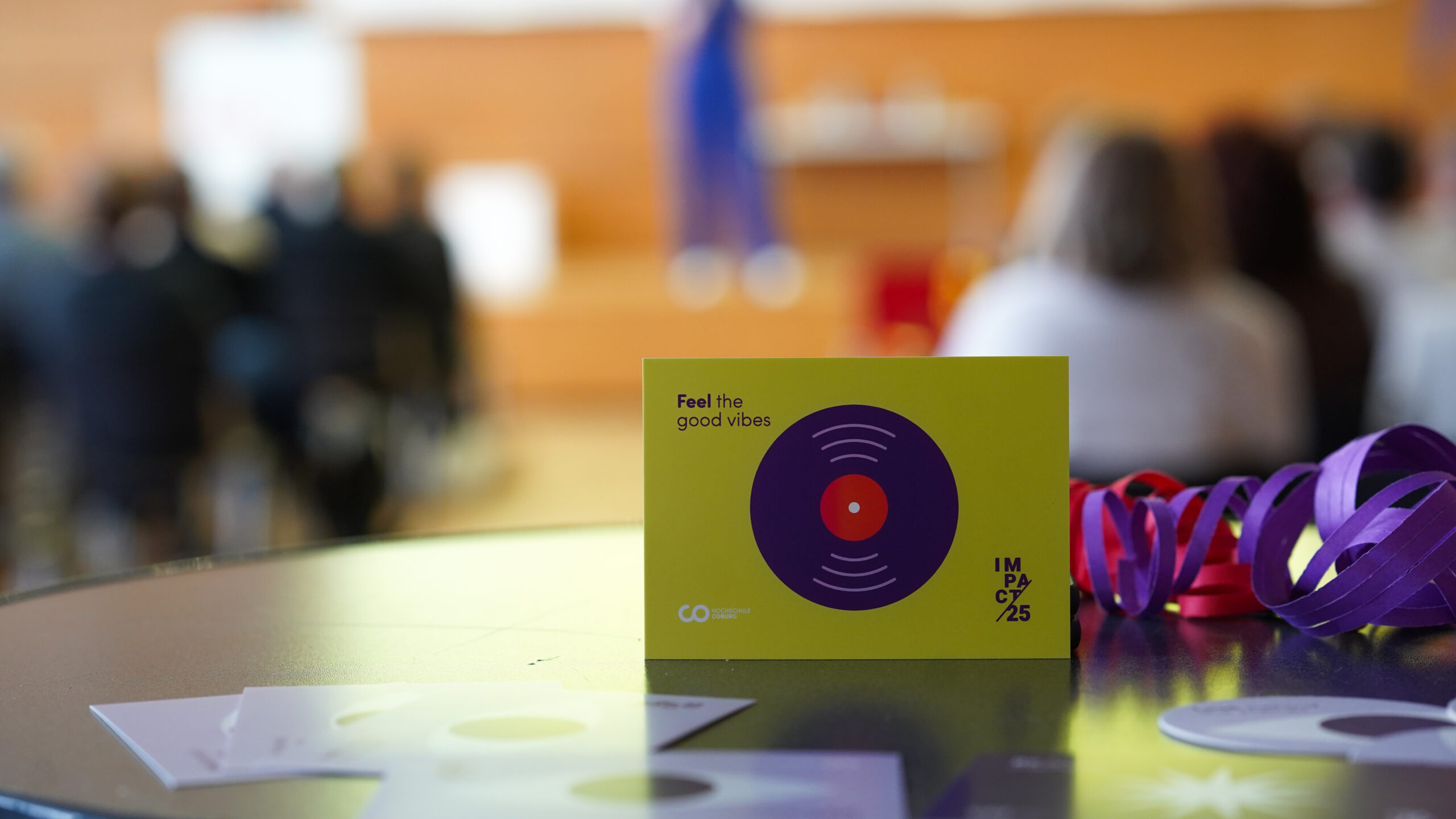 Eine Nahaufnahme einer Karte mit einem Schallplattenmotiv und dem Text "Feel the good vibes" auf einem Tisch. Bunte Bänder und unscharfe Menschen im Hintergrund evozieren eine Atmosphäre voller Kreativität und lebendigem Teamgeist bei einem Indoor-Treffen. at Hochschule Coburg