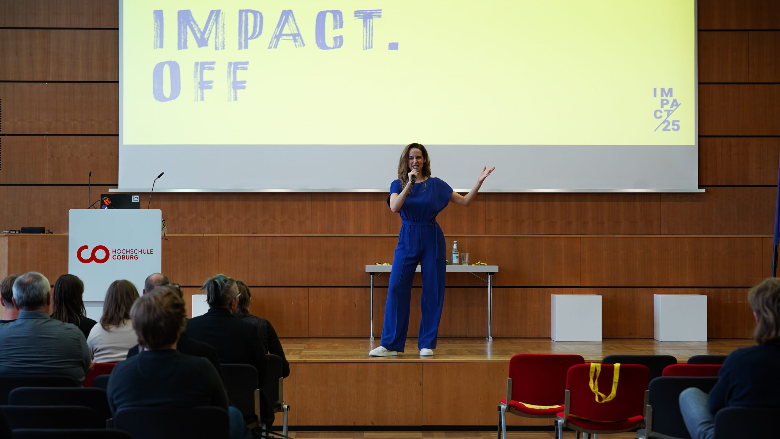 Eine Frau in einem blauen Overall spricht auf der Bühne mit einem Mikrofon und inspiriert das Publikum mit Kreativität. Hinter ihr erscheint auf einer großen Leinwand "IMPACT. OFF" und ein Podium mit der Aufschrift "Hochschule Coburg". at Hochschule Coburg