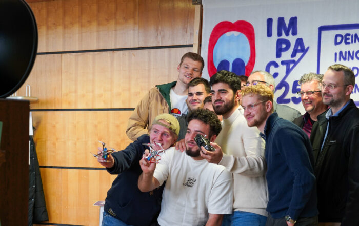 Eine Gruppe lächelnder Menschen posiert gemeinsam vor einer Holzwand und einem "Impact 25"-Banner mit der Aufschrift "Dein Innovationsfenster". Einige halten kleine Drohnen, auf denen Innovation und Teamgeist zu sehen sind, während sie sich für ein Gruppenfoto versammeln. at Hochschule Coburg