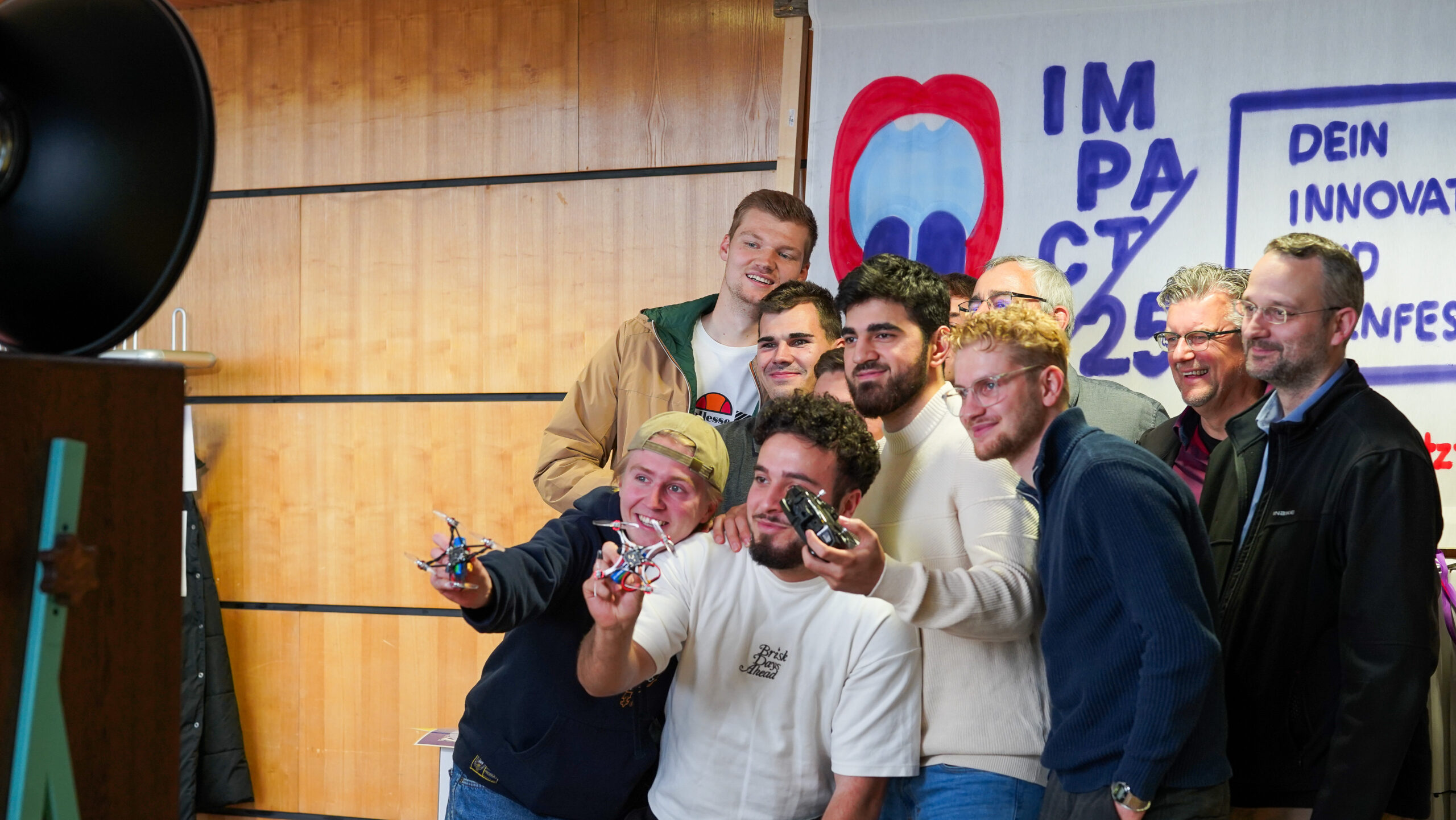 Eine Gruppe lächelnder Menschen posiert gemeinsam vor einer Holzwand und einem "Impact 25"-Banner mit der Aufschrift "Dein Innovationsfenster". Einige halten kleine Drohnen, auf denen Innovation und Teamgeist zu sehen sind, während sie sich für ein Gruppenfoto versammeln. at Hochschule Coburg