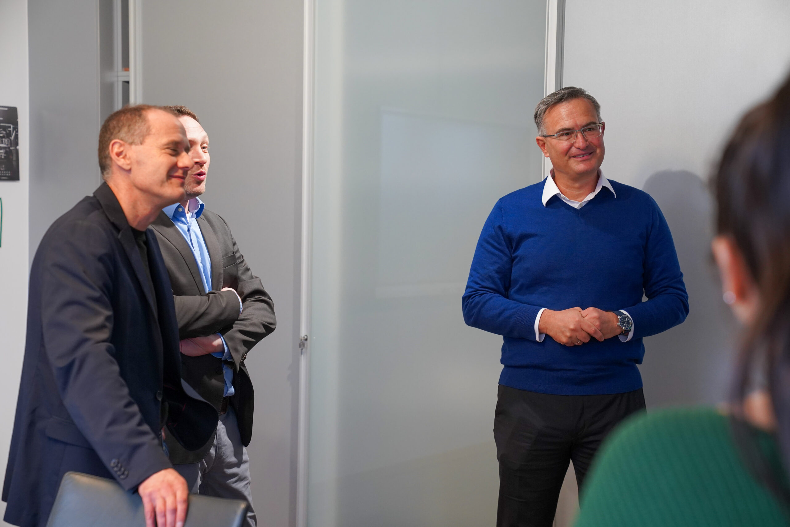 Drei Männer in Geschäftskleidung stehen und lächeln, während sie sich in einer modernen Büroumgebung über Projektmanagement unterhalten, wobei eine Person teilweise im Vordergrund zu sehen ist. at Hochschule Coburg