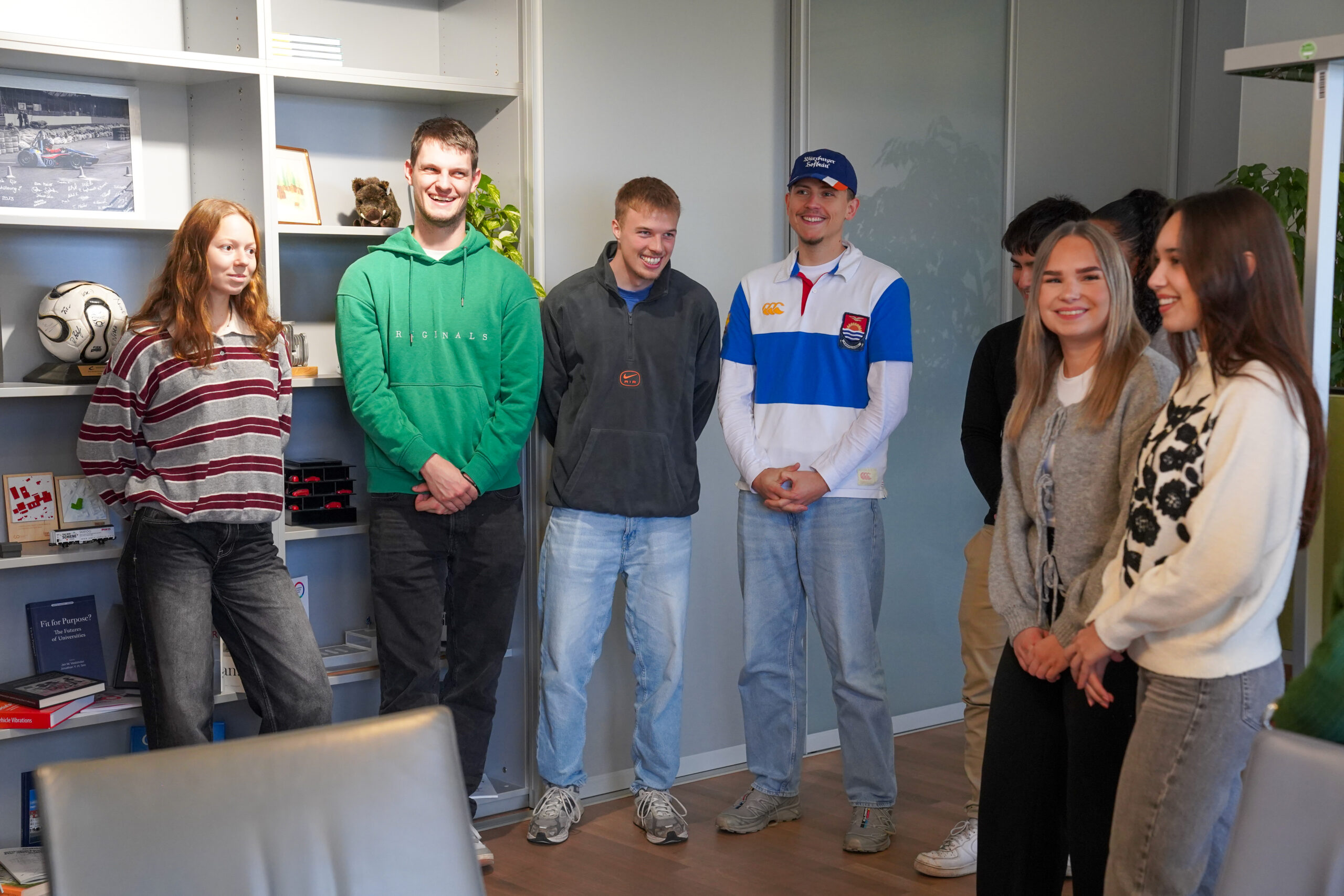 Eine Gruppe von sechs jungen Erwachsenen steht in einem Büro zusammen, lächelt und plaudert über ihre internationale Projektwoche. Im Hintergrund stehen Bücherregale mit Büchern, Dekorationen und einem Fußball. Die Stimmung wirkt freundlich und locker. at Hochschule Coburg