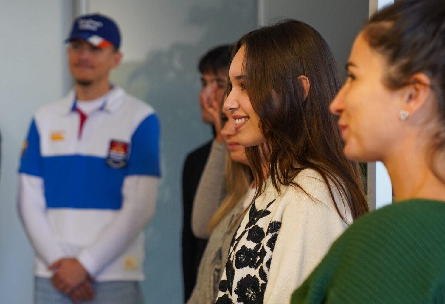 Eine Gruppe von sechs jungen Erwachsenen steht in einem Innenraum in einem Halbkreis, lächelt und scheint in ein Gespräch über eine internationale Projektwoche vertieft zu sein. Im Hintergrund sind Regale und Pflanzen zu sehen, die eine lockere und freundliche Atmosphäre schaffen. at Hochschule Coburg