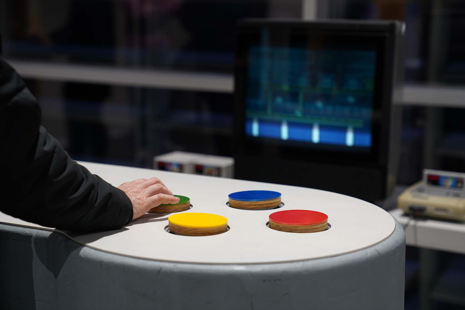 Eine Person drückt eine von vier großen, bunten Tasten - gelb, grün, blau und rot - auf einer ovalen Konsole an der Hochschule Coburg Informatik, während im Hintergrund ein Monitor KI-Grafiken anzeigt. at Hochschule Coburg