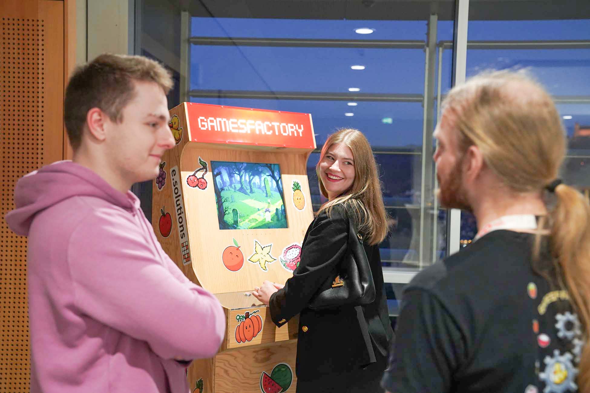 Drei Personen stehen neben einem hölzernen Spielautomaten mit der Aufschrift "GAMESFACTORY". Eine Person lächelt in die Kamera, während sie spielt, während die anderen sich unterhalten - und so den Informatik- und KI-Geist der Hochschule Coburg vor einer nächtlichen Fensterkulisse präsentieren. at Hochschule Coburg