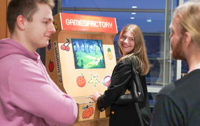 Drei Personen stehen um einen hölzernen Spielautomaten mit der Aufschrift "GAMESFACTORY". Einer spielt und lächelt in die Kamera, während sich die anderen in der Nähe unterhalten. Die Szene spielt sich in einem Innenraum der Hochschule Coburg ab, mit großen Fenstern und einer Atmosphäre von Informatik und KI-Innovation. at Hochschule Coburg