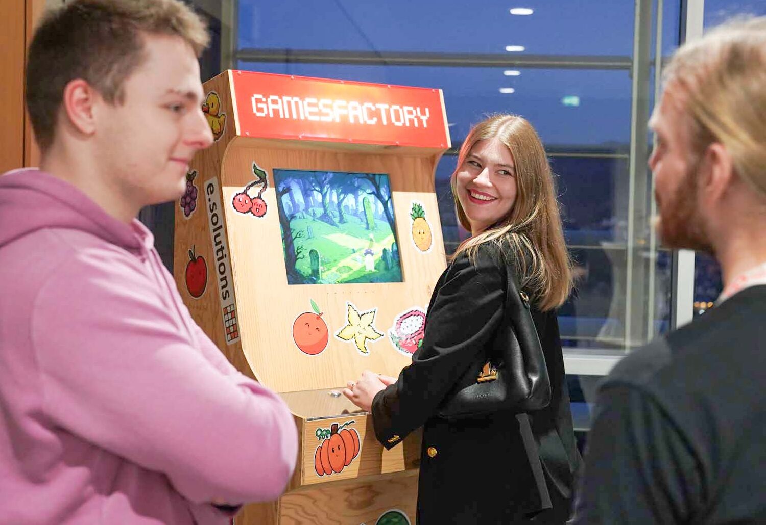 Drei Personen stehen um einen hölzernen Spielautomaten mit der Aufschrift "GAMESFACTORY". Einer spielt und lächelt in die Kamera, während sich die anderen in der Nähe unterhalten. Die Szene spielt sich in einem Innenraum der Hochschule Coburg ab, mit großen Fenstern und einer Atmosphäre von Informatik und KI-Innovation. at Hochschule Coburg
