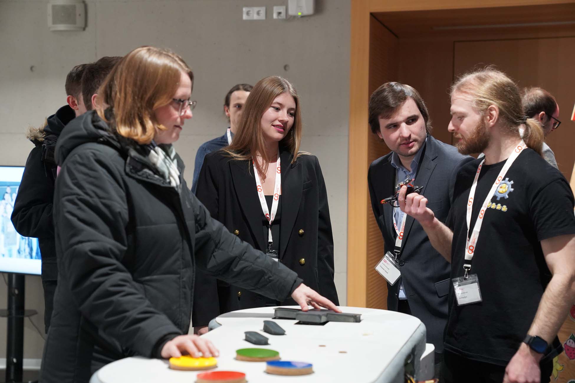 Eine Gruppe von Menschen steht um einen weißen Tisch mit großen farbigen Knöpfen an der Hochschule Coburg. Eine Person in einer schwarzen Jacke drückt einen Knopf, während eine andere, möglicherweise aus der Informatik, etwas über KI erklärt, während die anderen drinnen aufmerksam zusehen und zuhören. at Hochschule Coburg