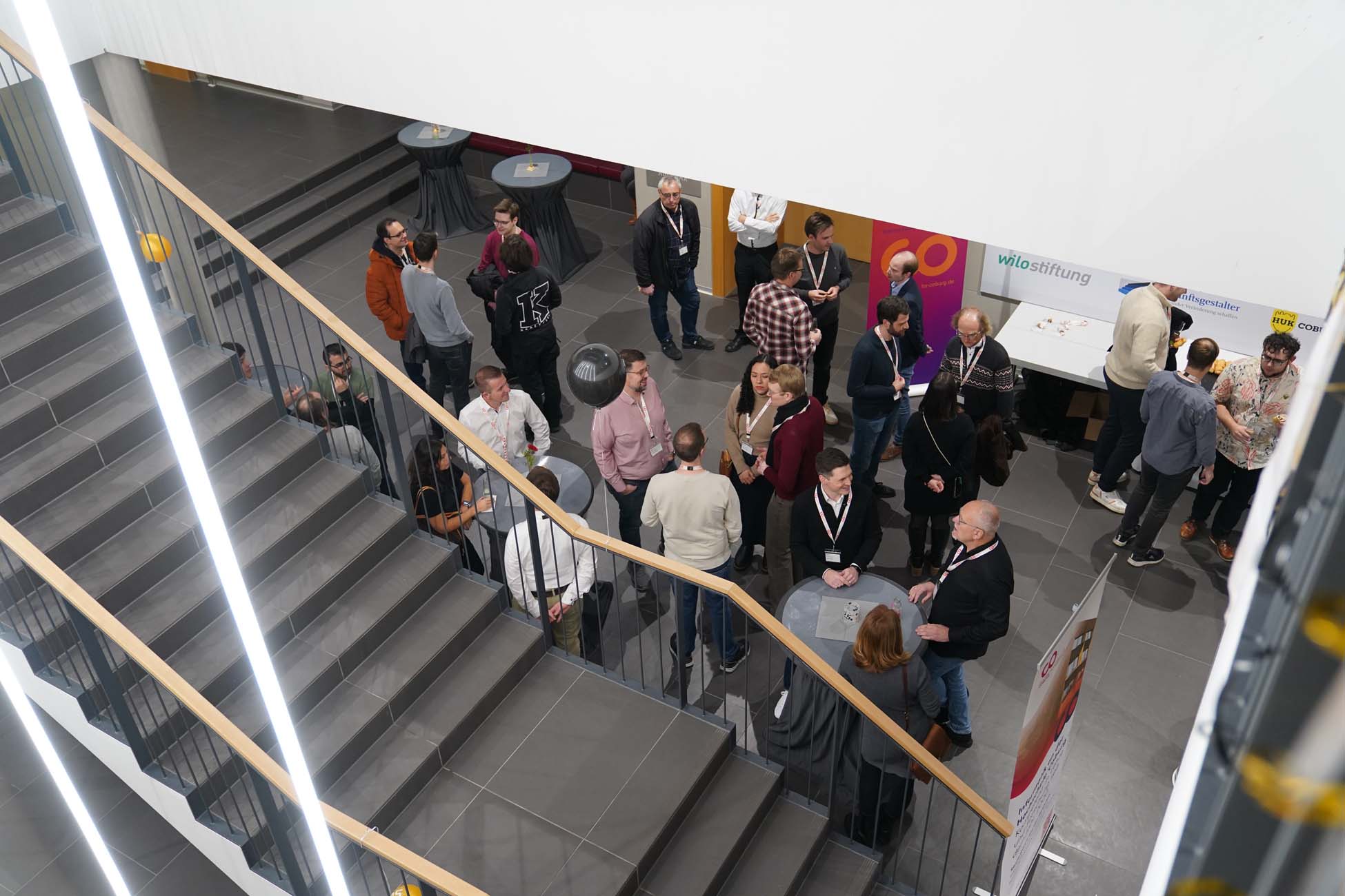 Eine Gruppe von Personen trifft sich an Stehtischen in einem modernen Atrium der Hochschule Coburg, von oben gesehen in der Nähe einer Treppe, während einer offenbar beruflichen oder akademischen Veranstaltung zum Thema KI und Informatik. at Hochschule Coburg