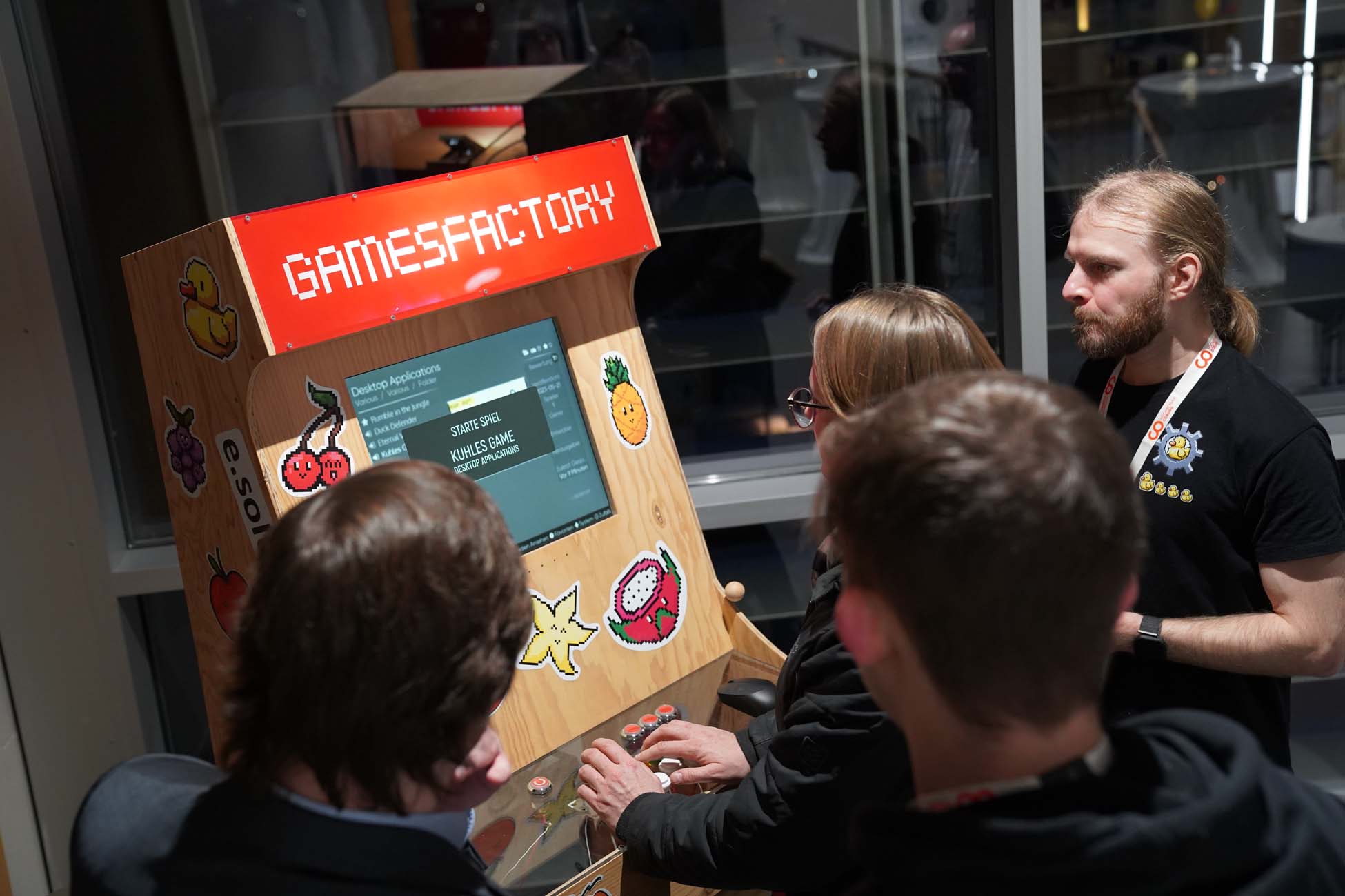 Vier Personen stehen um einen hölzernen Spielautomaten mit der Aufschrift "GAMESFACTORY" an der Hochschule Coburg und interagieren mit den Bedienelementen und dem Bildschirm. Ein Mann scheint KI-Konzepte zu erklären, während die anderen spielen. Der Bereich ist gut beleuchtet und befindet sich in einem Innenraum. at Hochschule Coburg
