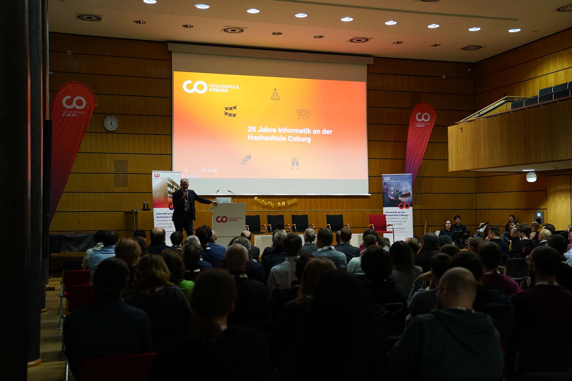 Ein Redner wendet sich an das Publikum in einem Hörsaal mit einer großen orangefarbenen Leinwand, auf der "25 Jahre Informatik an der Hochschule Coburg" steht. Die Veranstaltung hebt die Errungenschaften der Informatik und der KI hervor, mit Bannern, die auf beiden Seiten der Bühne zu sehen sind. at Hochschule Coburg