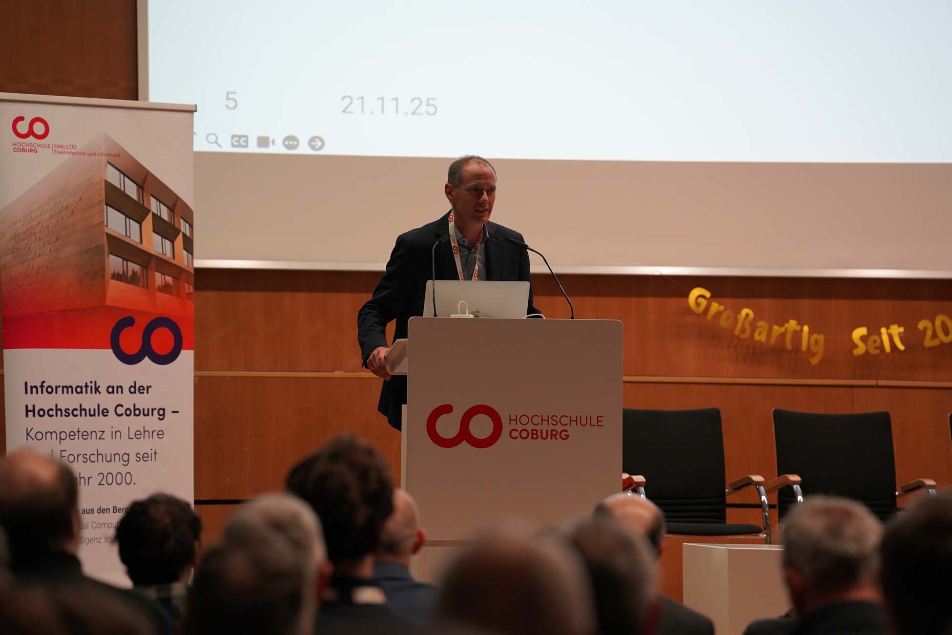 Ein Mann steht an einem Podium und hält einen Vortrag über KI an der Hochschule Coburg. Zu sehen sind sitzende Zuhörer, im Hintergrund ein Banner und ein Informationsplakat über Informatik an der Hochschule Coburg. at Hochschule Coburg