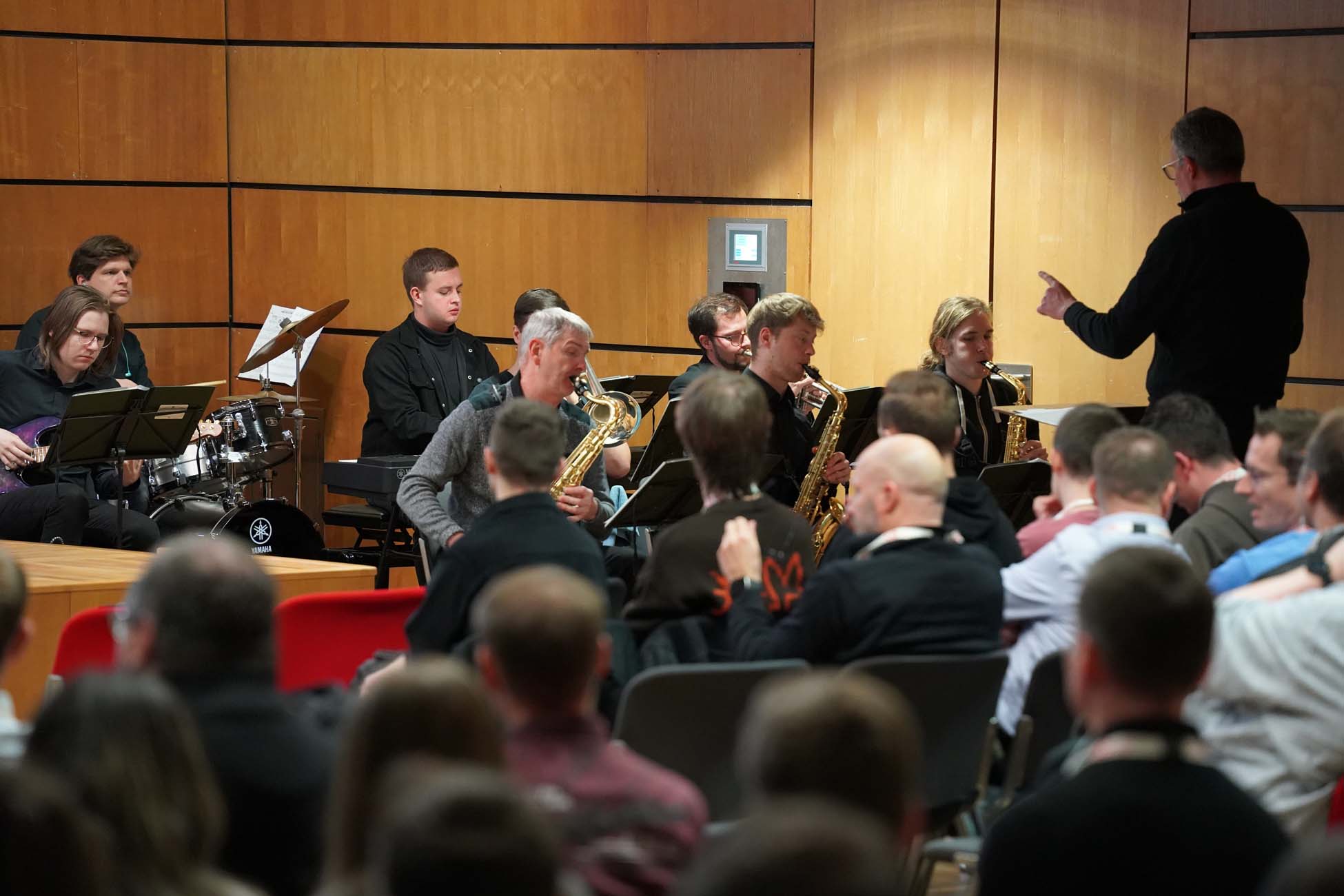 In einem holzgetäfelten Raum der Hochschule Coburg, in dem auch KI- und Informatikstudenten die Kunst der Live-Musik zu schätzen wissen, steht eine Jazzband unter der Leitung eines Dirigenten auf der Bühne, während das Publikum aufmerksam zusieht. at Hochschule Coburg