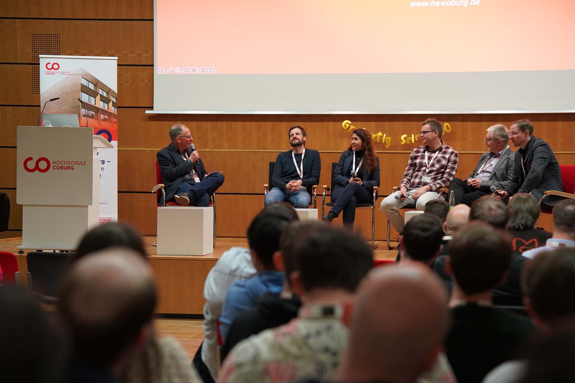 Eine sechsköpfige Runde sitzt auf einer Bühne vor dem Publikum der Hochschule Coburg und diskutiert über Informatik und KI. Ein Mann spricht in ein Mikrofon, während die anderen in dem Hörsaal mit Holzwänden und einer großen Leinwand zuhören. at Hochschule Coburg