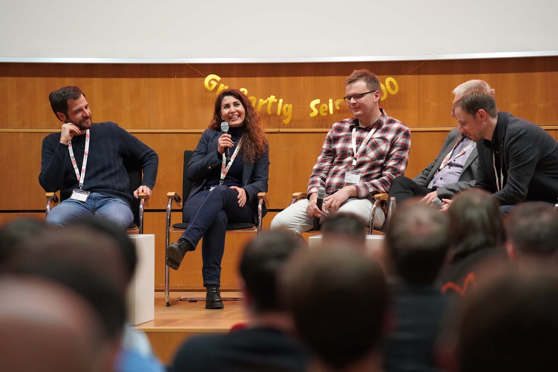 Fünf Personen sitzen auf der Bühne vor einem Publikum an der Hochschule Coburg und führen eine Podiumsdiskussion über KI. Die Frau in der Mitte spricht in ein Mikrofon, während die anderen zuhören und lächeln. Hinter ihnen hängt ein gelbes Transparent. at Hochschule Coburg