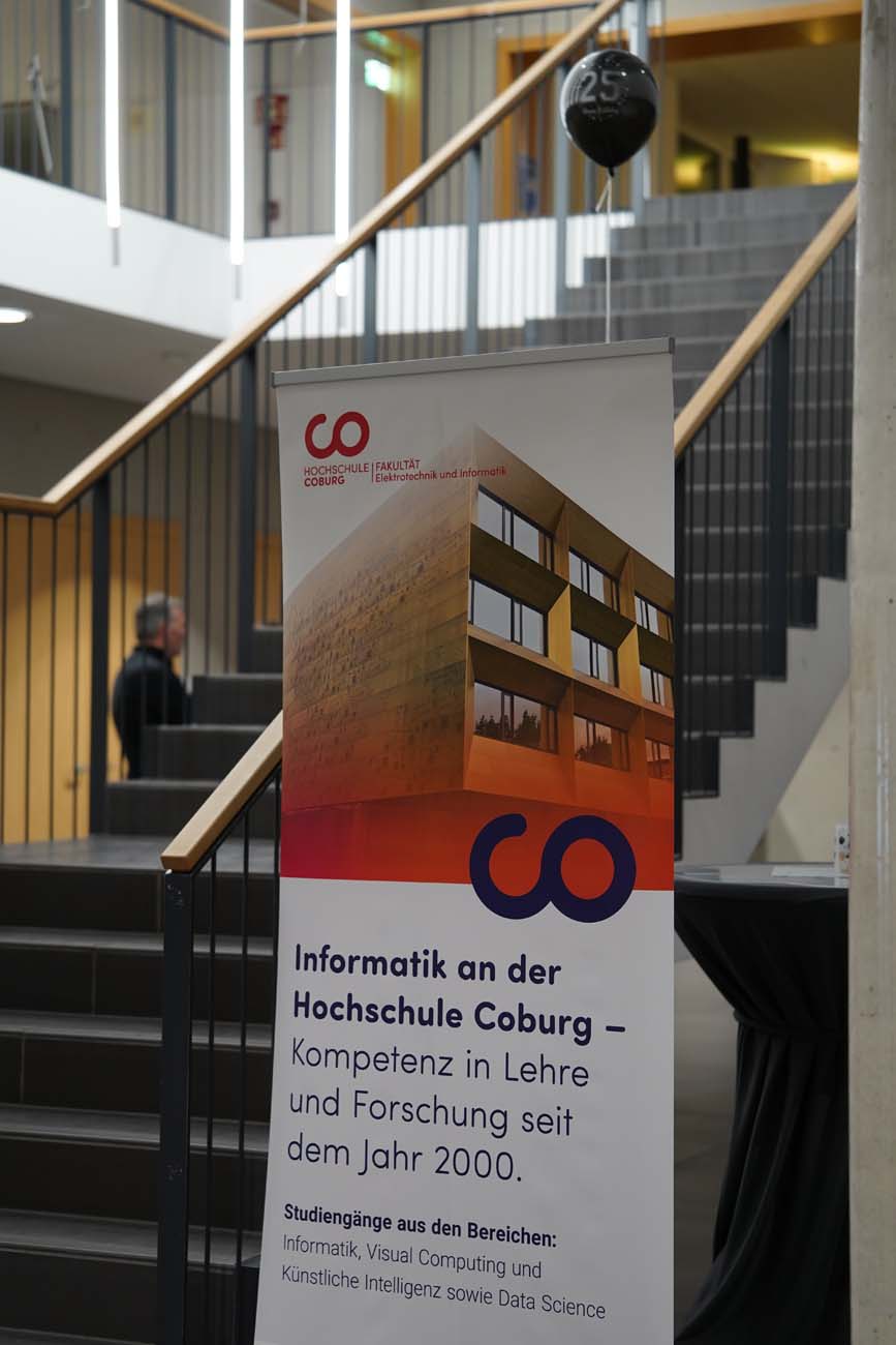 Vor einem Treppenhaus steht ein Banner, das für Informatik und KI an der Hochschule Coburg wirbt. Es hebt die Kompetenz in Lehre und Forschung seit 2000 hervor und listet verwandte Studienrichtungen auf. Darüber hängt ein schwarzer Luftballon mit der Zahl "25". at Hochschule Coburg