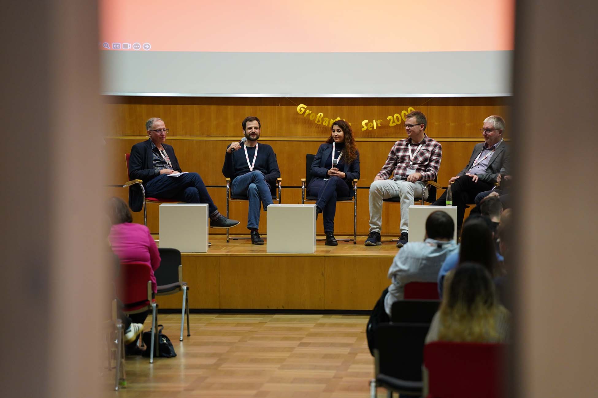 Bei einer Podiumsdiskussion zum Thema Informatik an der Hochschule Coburg sitzen fünf Personen mit Mikrofon und Schlüsselband auf einer Bühne und wenden sich an das Publikum in einem Hörsaal. Die Veranstaltung wirkt förmlich, über den Podiumsteilnehmern hängt eine orangefarbene Leinwand. at Hochschule Coburg