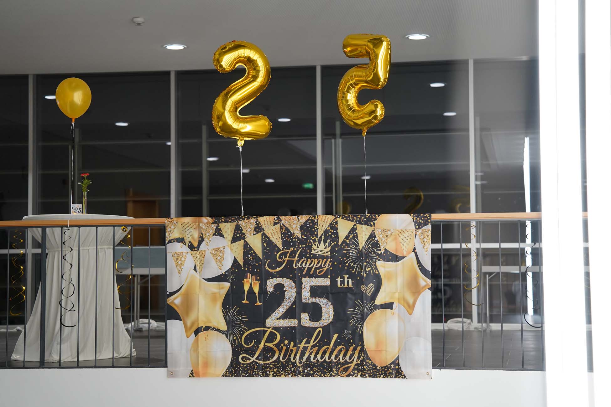 Ein dekorierter Bereich mit goldenen Luftballons in Form von "2" und "5", ein Banner mit der Aufschrift "Happy 25th Birthday", goldene und weiße Luftballons und ein kleiner Tisch mit einem gelben Luftballon und einer einzelnen Blume in einer Vase feiern das KI an der Hochschule Coburg. at Hochschule Coburg