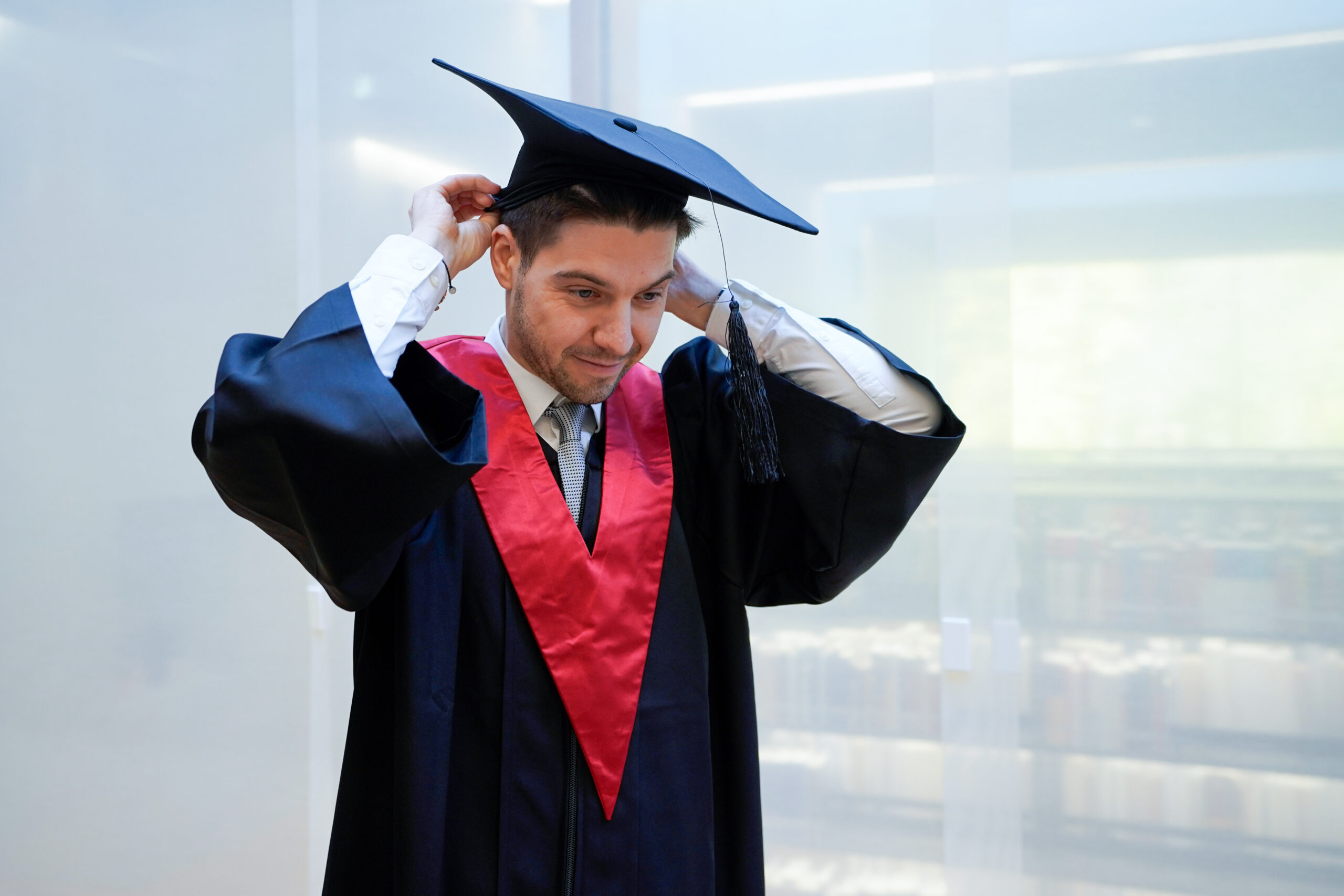 Ein Mann in Talar und Mütze rückt seine Quaste zurecht und lächelt leicht - bereit, die Zukunft zu gestalten. Im unscharfen Hintergrund sind Regale mit Büchern zu sehen. at Hochschule Coburg