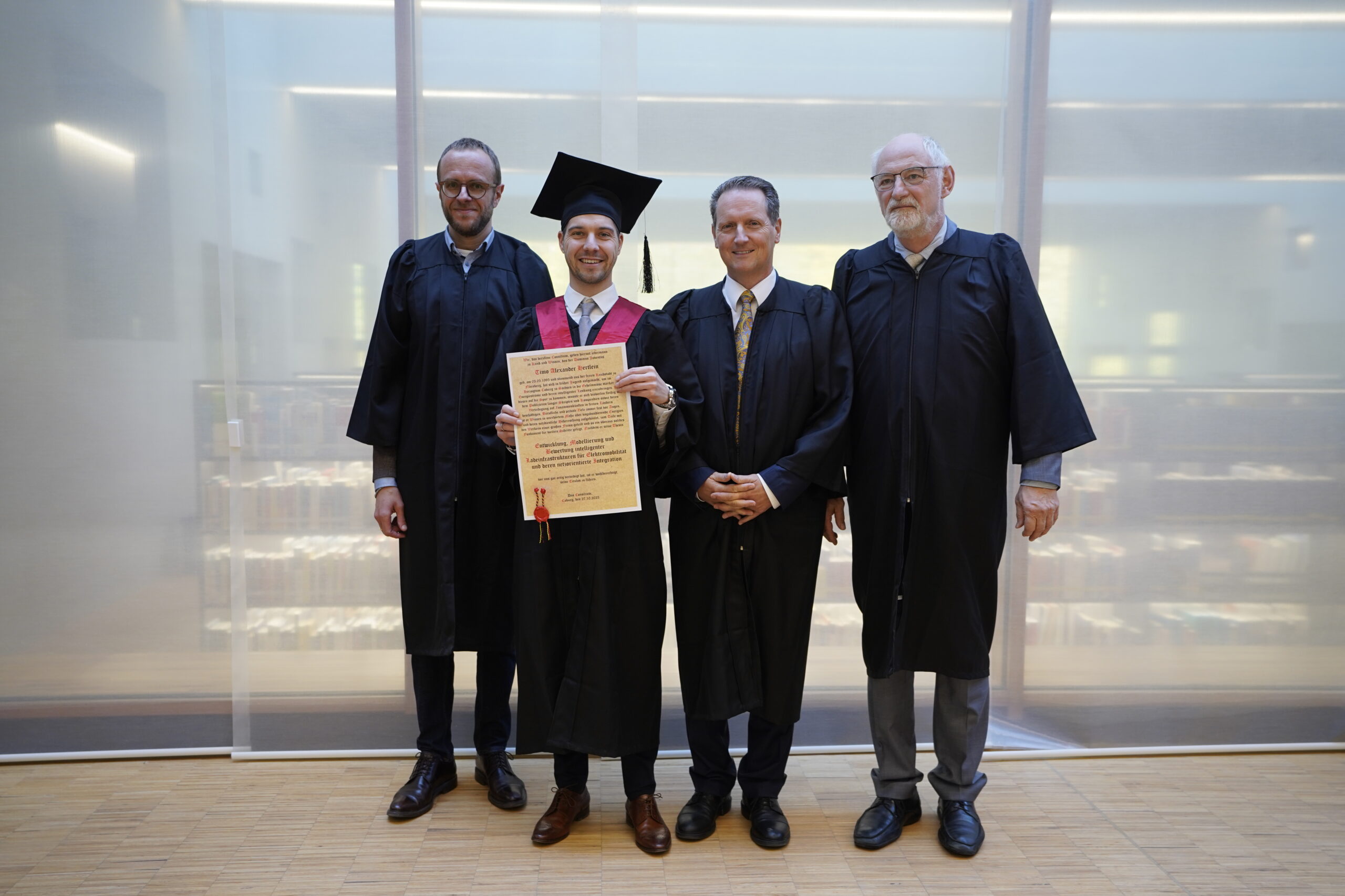 Vier Männer stehen in einem Innenraum; einer trägt eine Abschlusskappe und einen Talar und hält eine Urkunde in der Hand, während die anderen in akademischen Roben neben ihm lächeln und vor einem modernen, lichtdurchfluteten Hintergrund ihre Erfolge in Elektromobilität feiern. at Hochschule Coburg