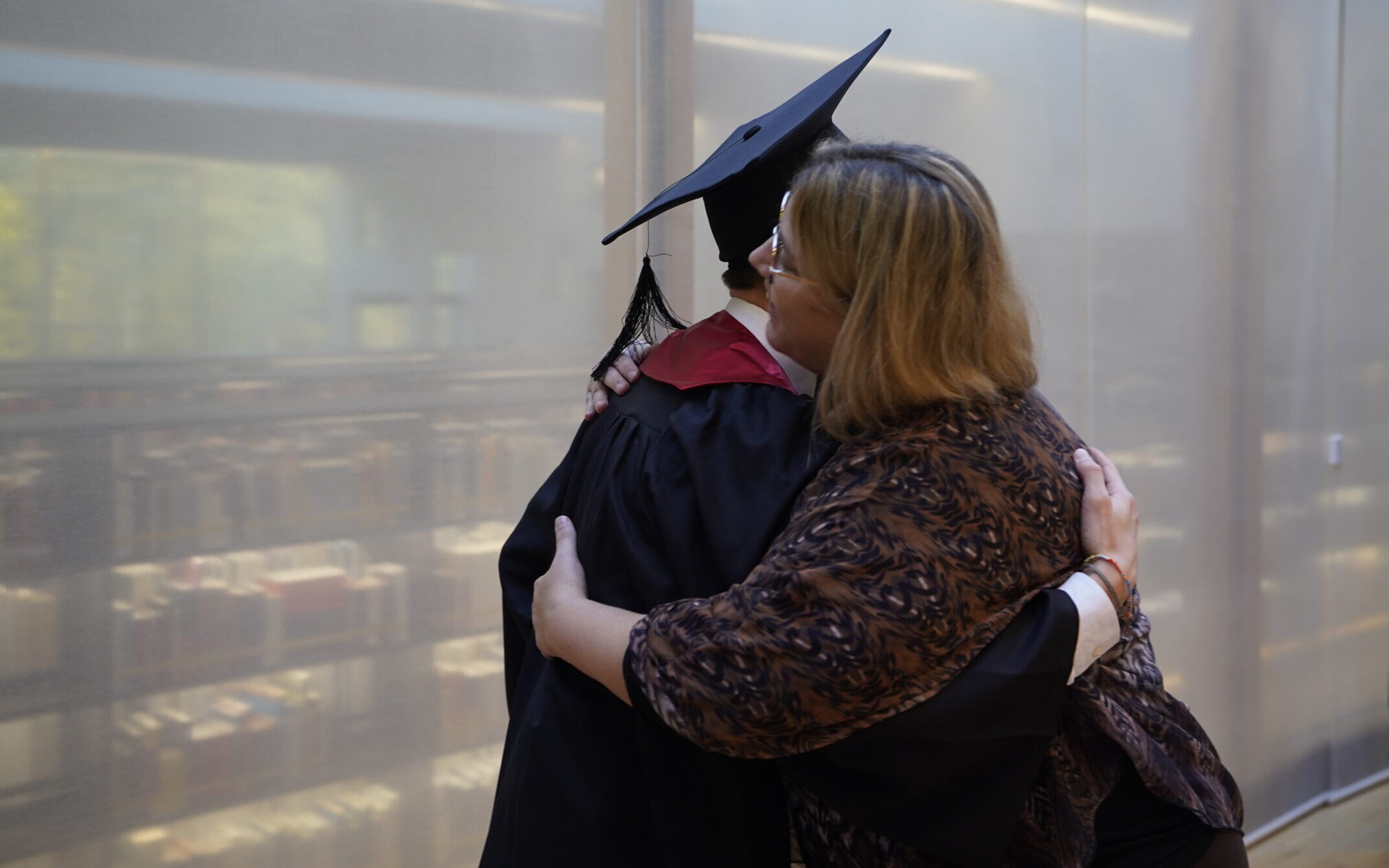 Ein Hochschulabsolvent mit Kappe und Talar umarmt eine Frau mit hellbraunem Haar und Brille in einem Innenraum und feiert eine Zukunft voller Möglichkeiten, während andere Menschen im Hintergrund stehen und gehen. at Hochschule Coburg